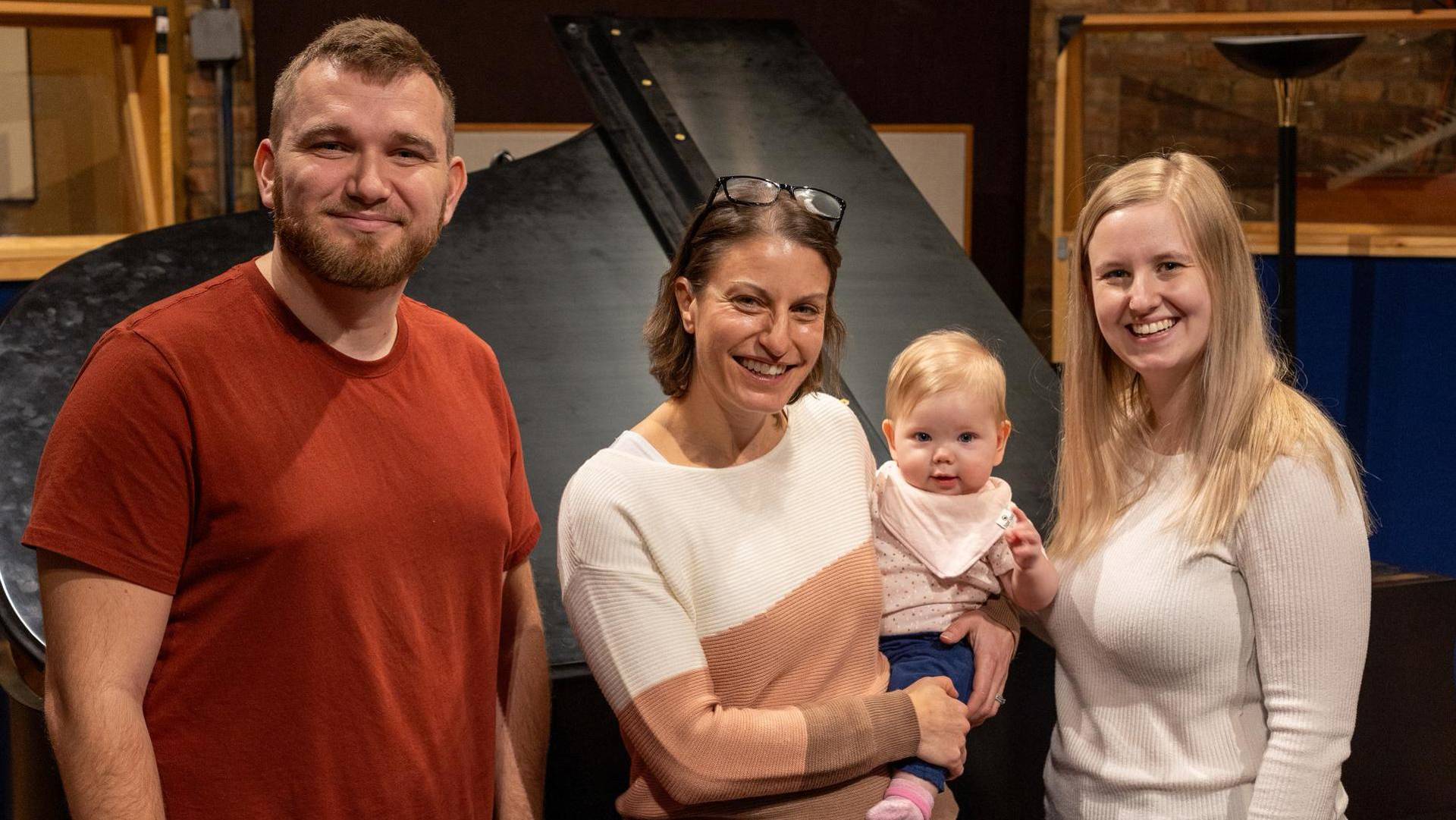 A man, two women and a baby posing together for a picture. They all have light complexion and are smiling.