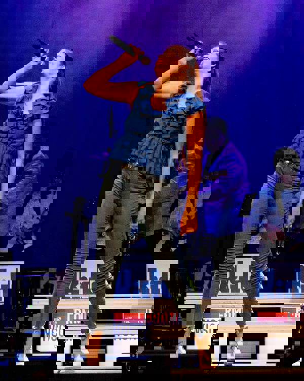 A young performer with light complexion, brown hair, a blue top and blue jeans singing into a microphone.