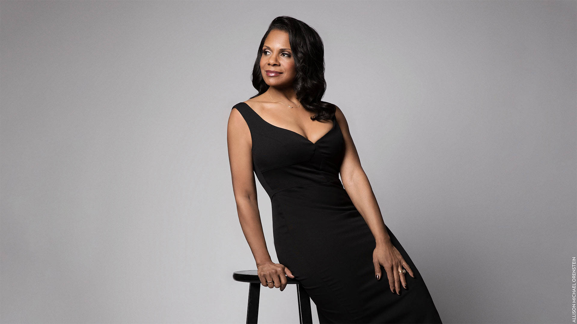 Audra McDonald, a woman with dark brown hair and a dark complexion, wears a black dress and sits on a stool.