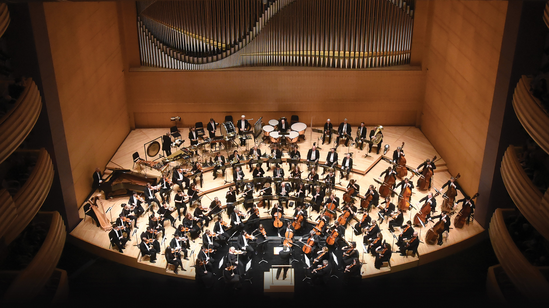 An aerial view of a symphony orchestra on stage. Large pipes from an organ appear behind them.