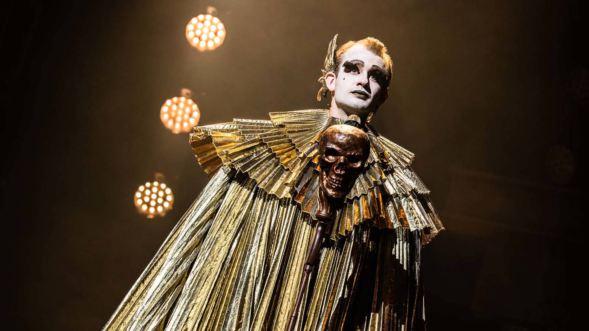 A caucasian man wearing white face make-up and lashes stands wearing a shiny gold pleated robe with a gold skull in the center.