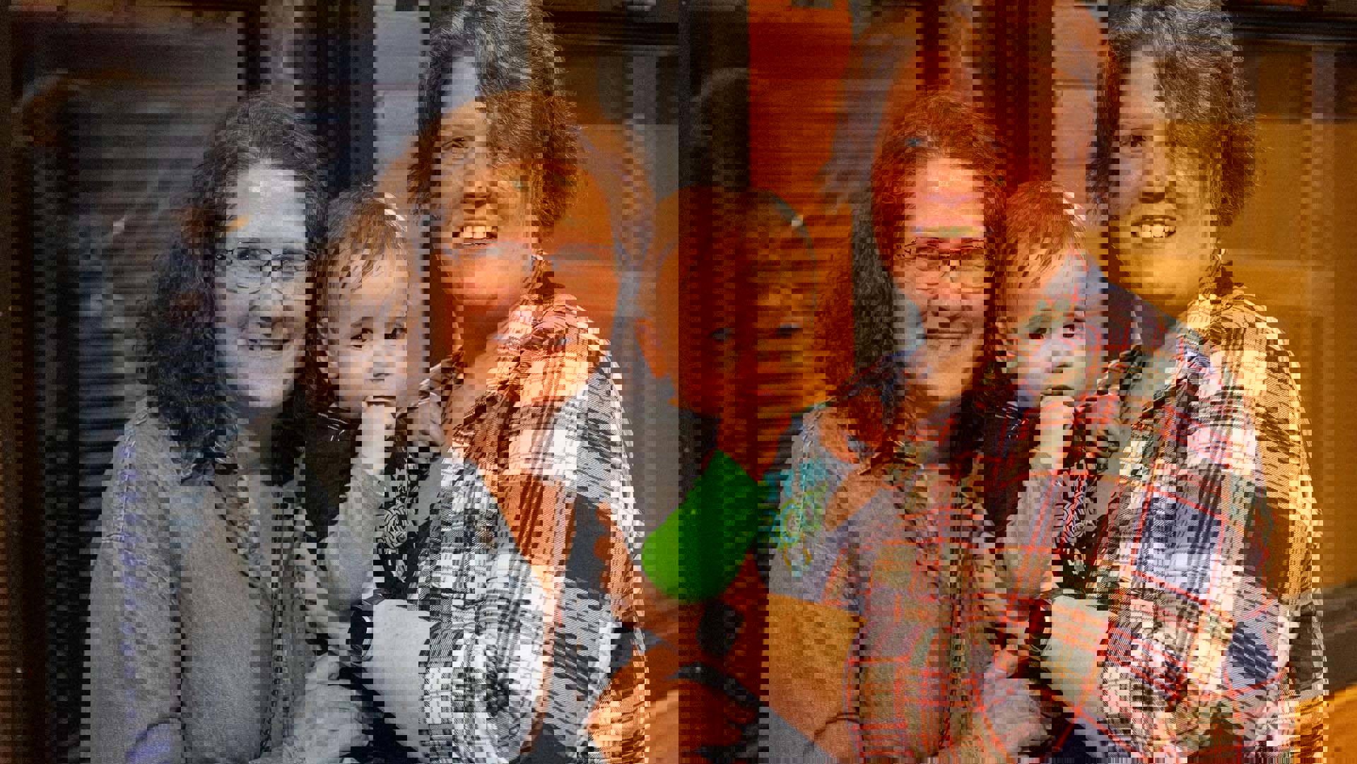 Two women with light complexion and brown hair holding a baby boy in a blue and green shirt.