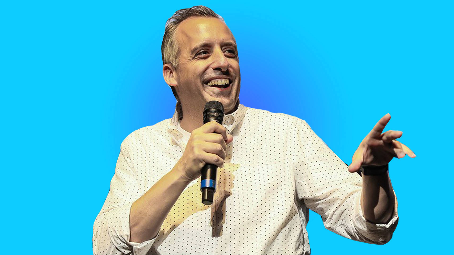 A middle-aged man smiling and holding a microphone. He is wearing a white button-down shirt and standing in front of a blue background.