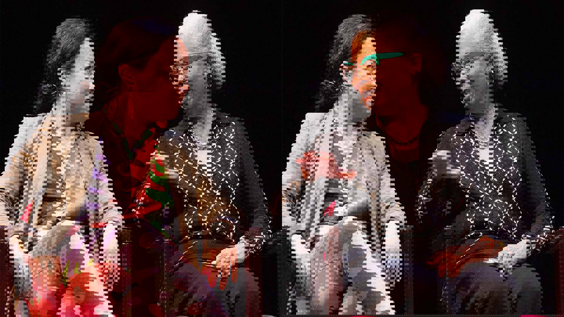 An asian woman with dark hair and colorful clothing sits next to an older african american woman with grey hair and a dark outfit. They are talking to eachother.