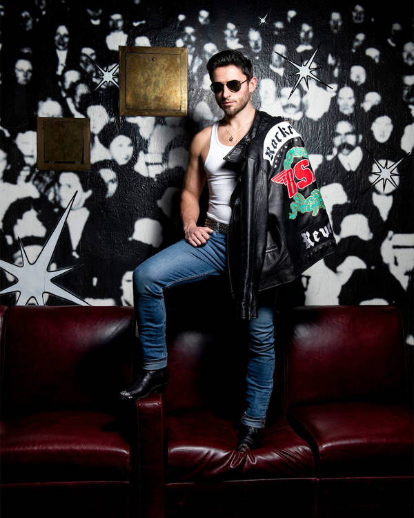 A man with short brown hair and sunglasses wearing jeans and white tank top with a leather jack over his shoulder. He is standing on a red leather couch.