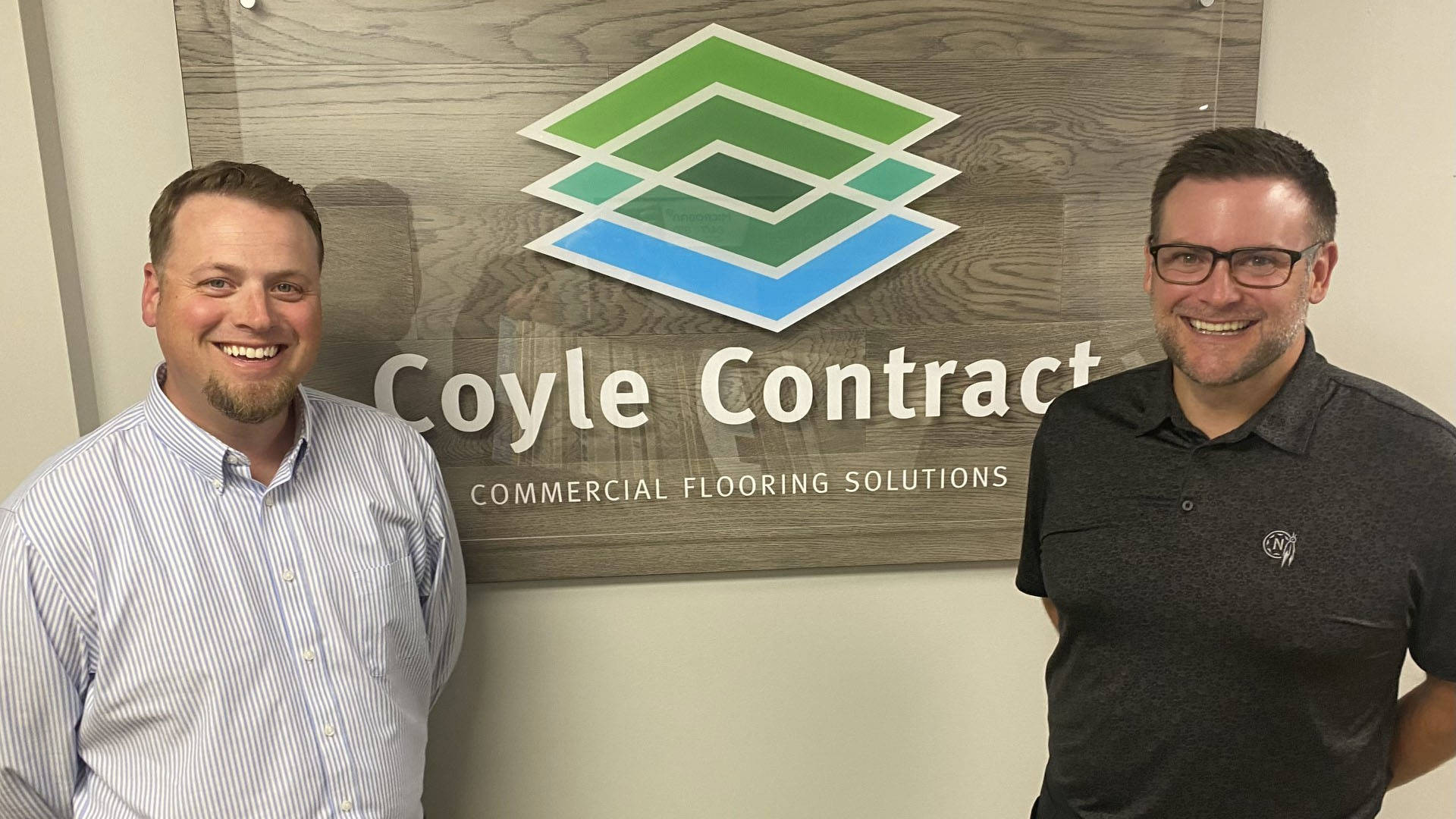 Two smiling caucasian men with short brown hair stand for a photo in front of a green blue and white company logo. The logo reads: Coyle Contract Commercial Flooring Solutions"