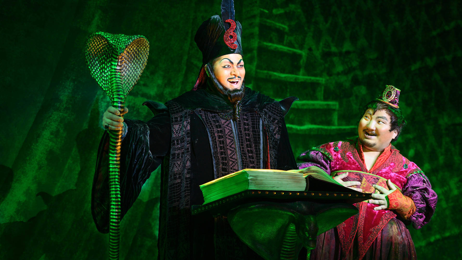 Two sinister looking men standing in front of a dark green staircase. The man on the left is dressed in black robes and holding a snake staff. He is standing in front of a podium with a large book opened. To his right the other man is wearing a pink robe and laughing.