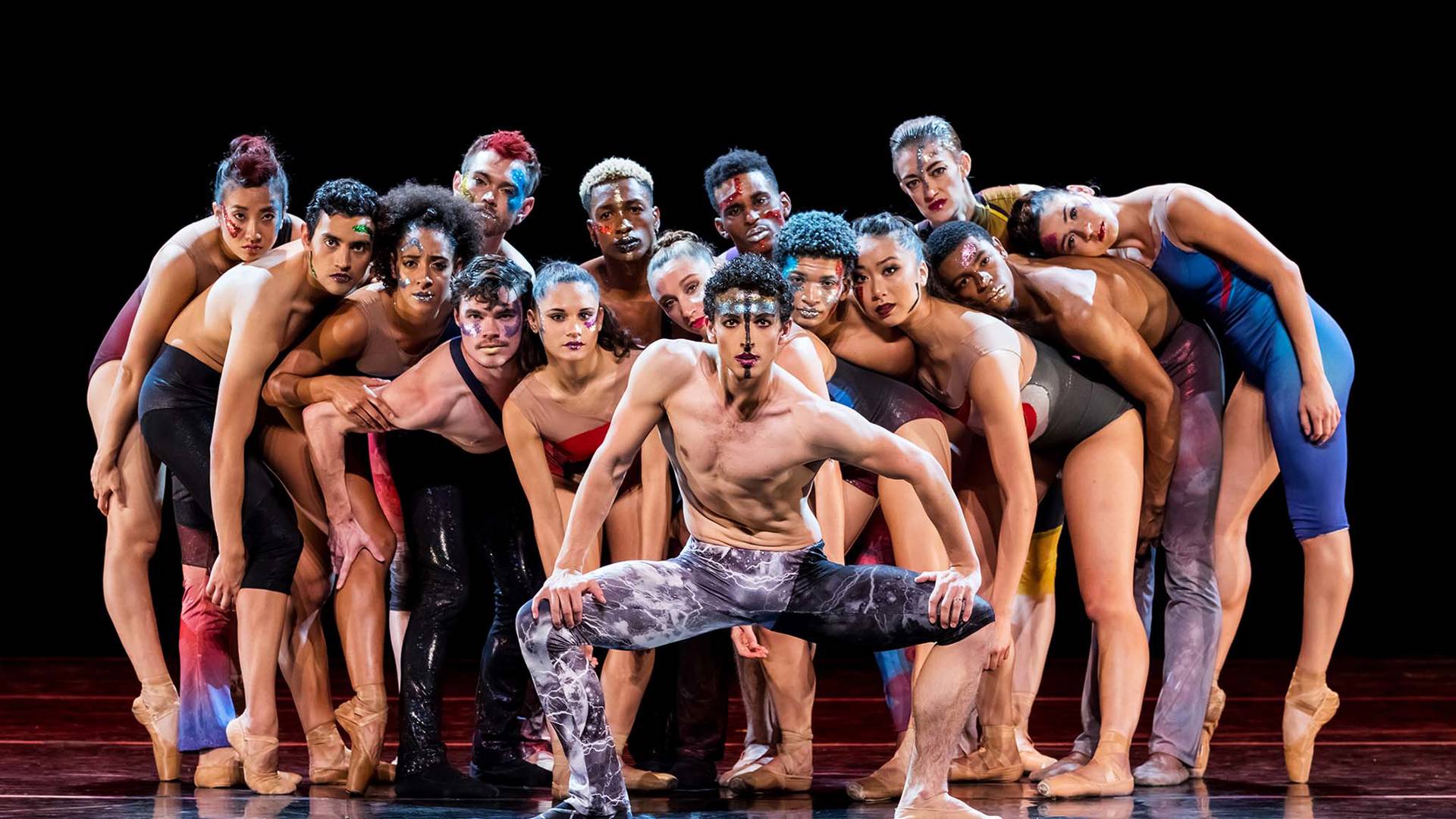 A group of diverse dancers in red and blue minimal skin-tight clothing crowding around each other looking forward.