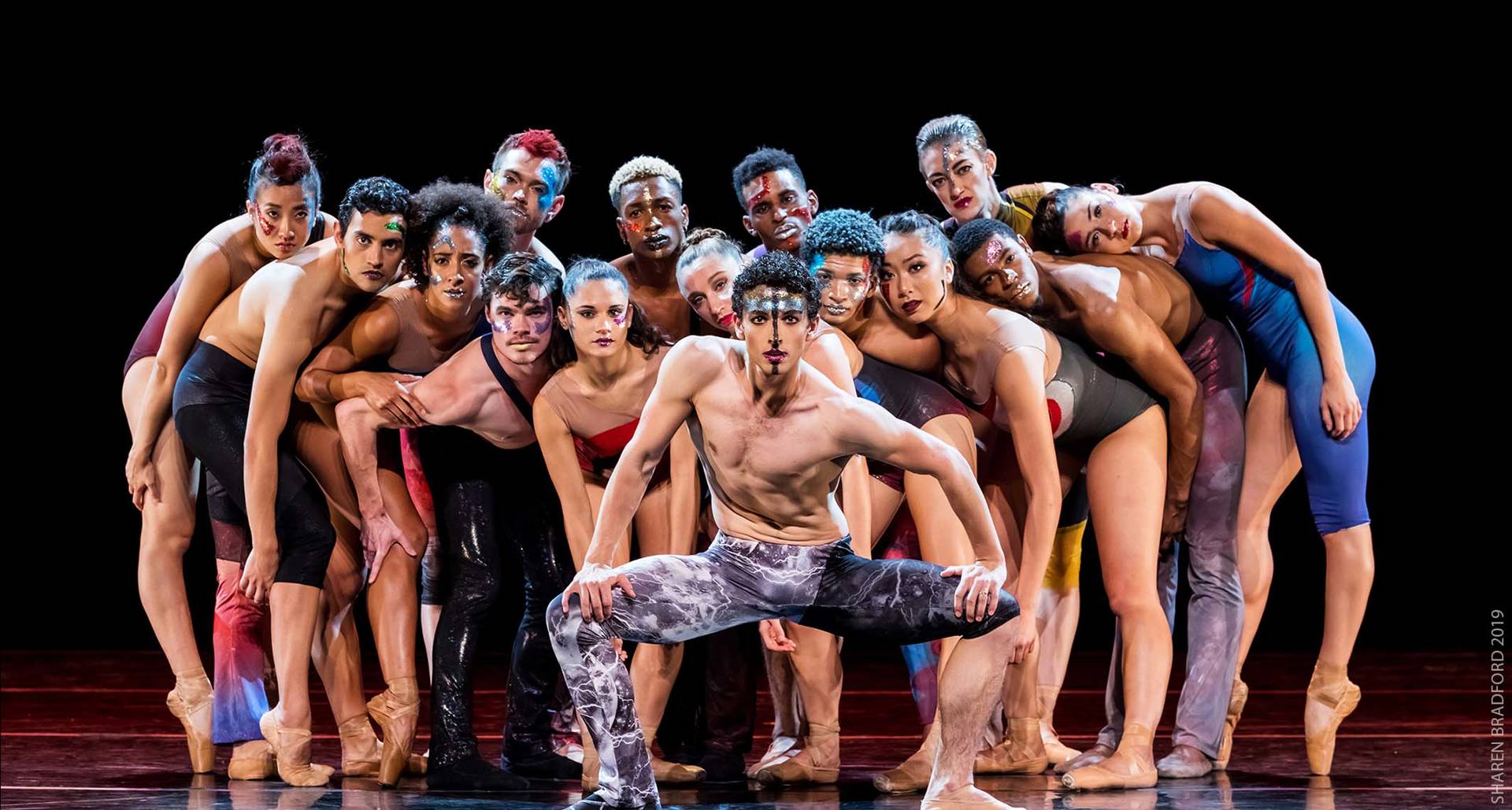 A group of diverse dancers in red and blue minimal skin-tight clothing crowding around each other looking forward.