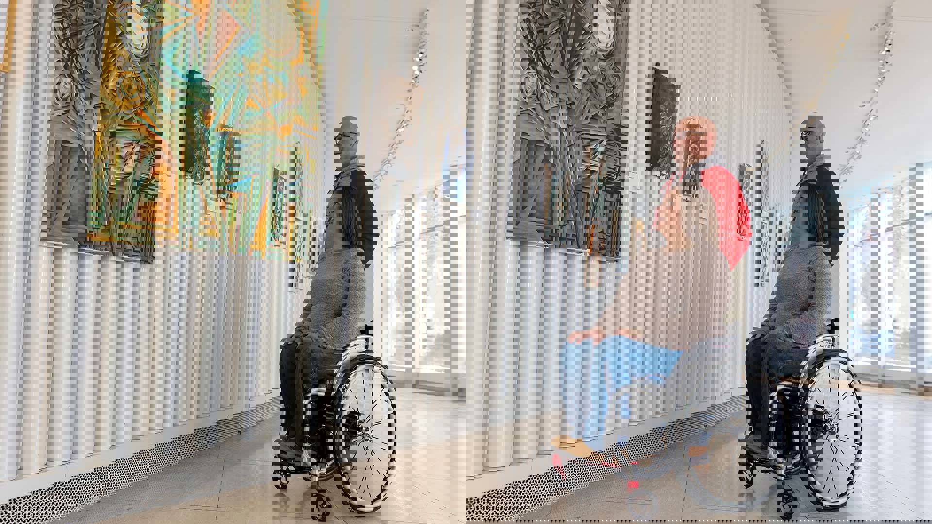 A woman with light complexion and brown hair sits in a wheelchair and looks at a large painting in an art exhibit with a man in a red and grey sweatshirt. sweatshirt.