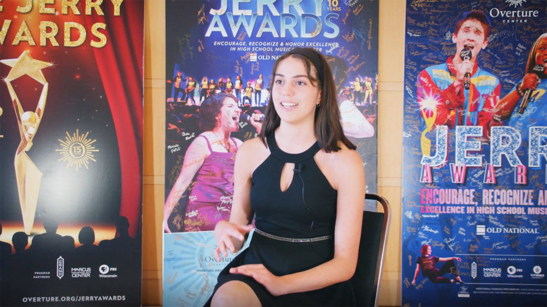 A teenage girl with medium length brown hair and light complexion, wearing a black dress, sitting in a black chair in front of three large "Jerry Awards" posters.