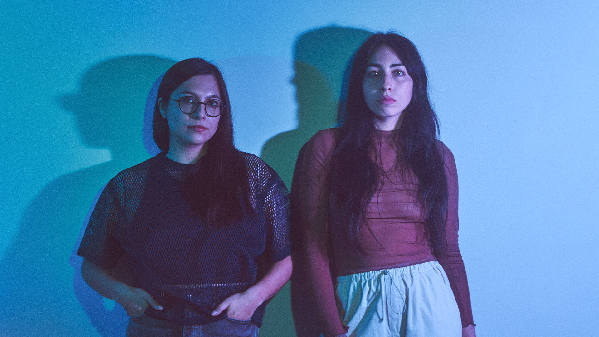 Two women with light complexion and dark hair standing against a blue wall. The woman on the left has a dark outfit and glasses. The woman on the right has a red top and tan pants. Their shadows are cast on the wall.