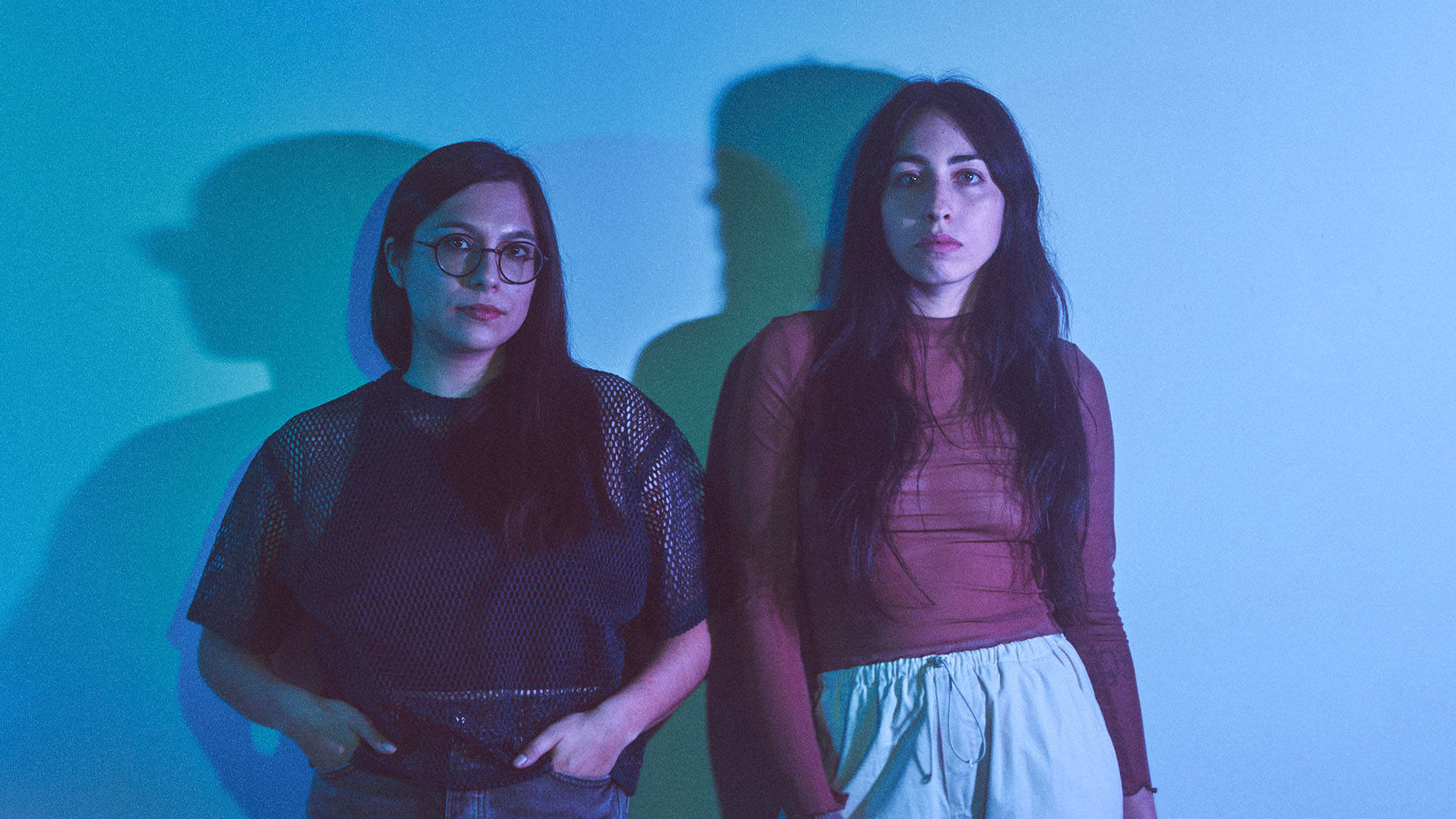 Two women with light complexion and dark hair standing against a blue wall. The woman on the left has a dark outfit and glasses. The woman on the right has a red top and tan pants. Their shadows are cast on the wall.