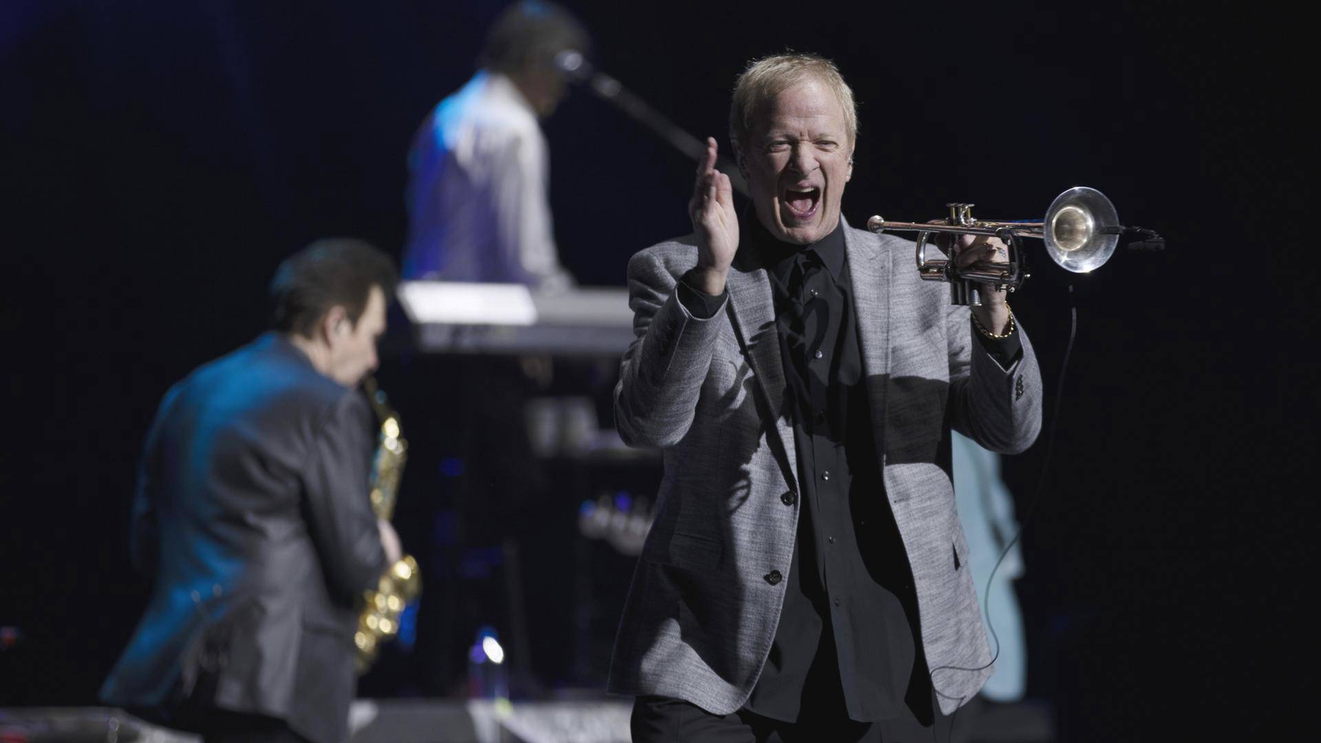 A man in a gray jacket holding a trumpet and yelling excitedly.