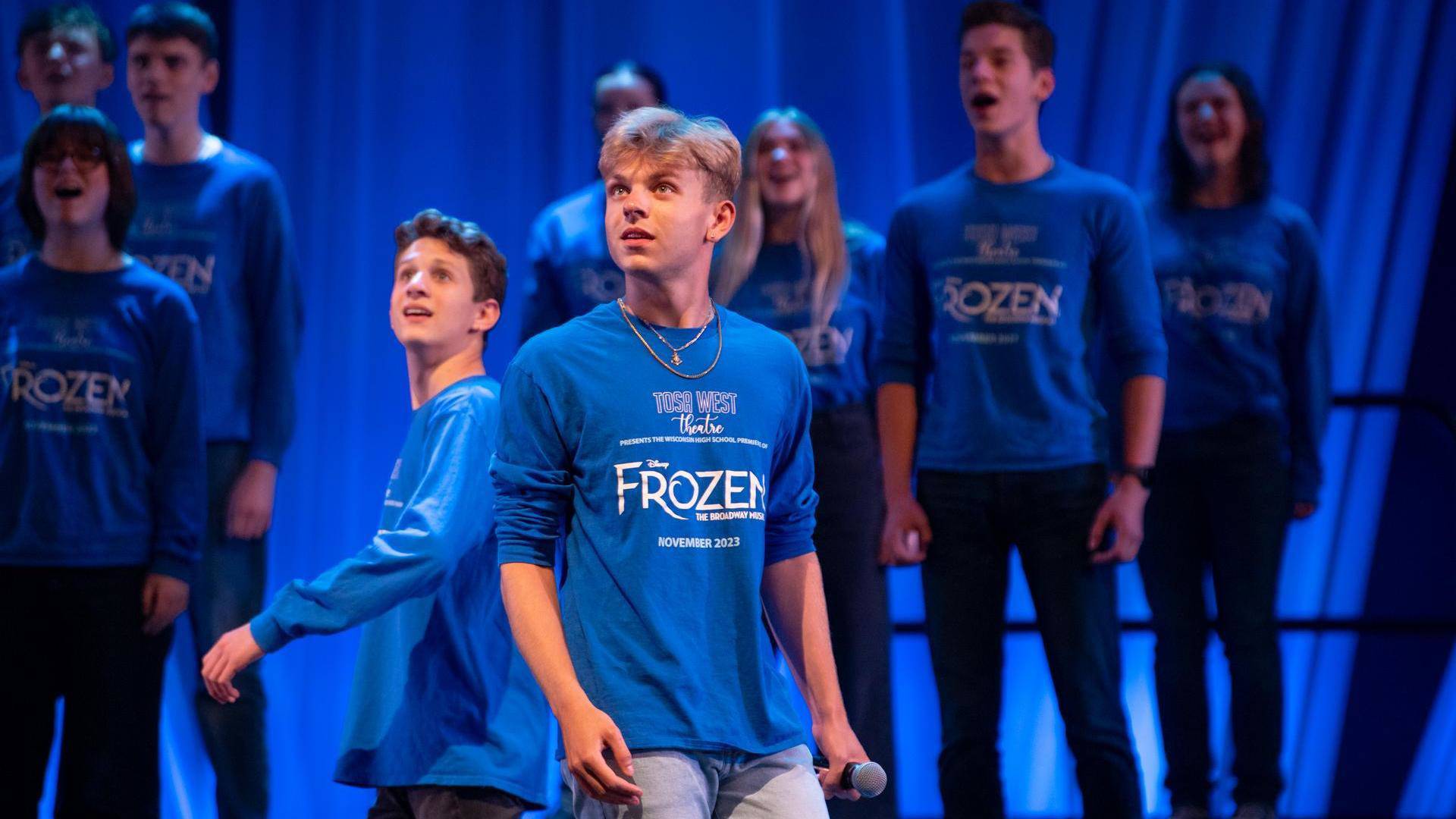 A high school aged performer in a blue t shirt with blond hair and light complexion. Other students in matching shirts are behind him. The background is also blue.