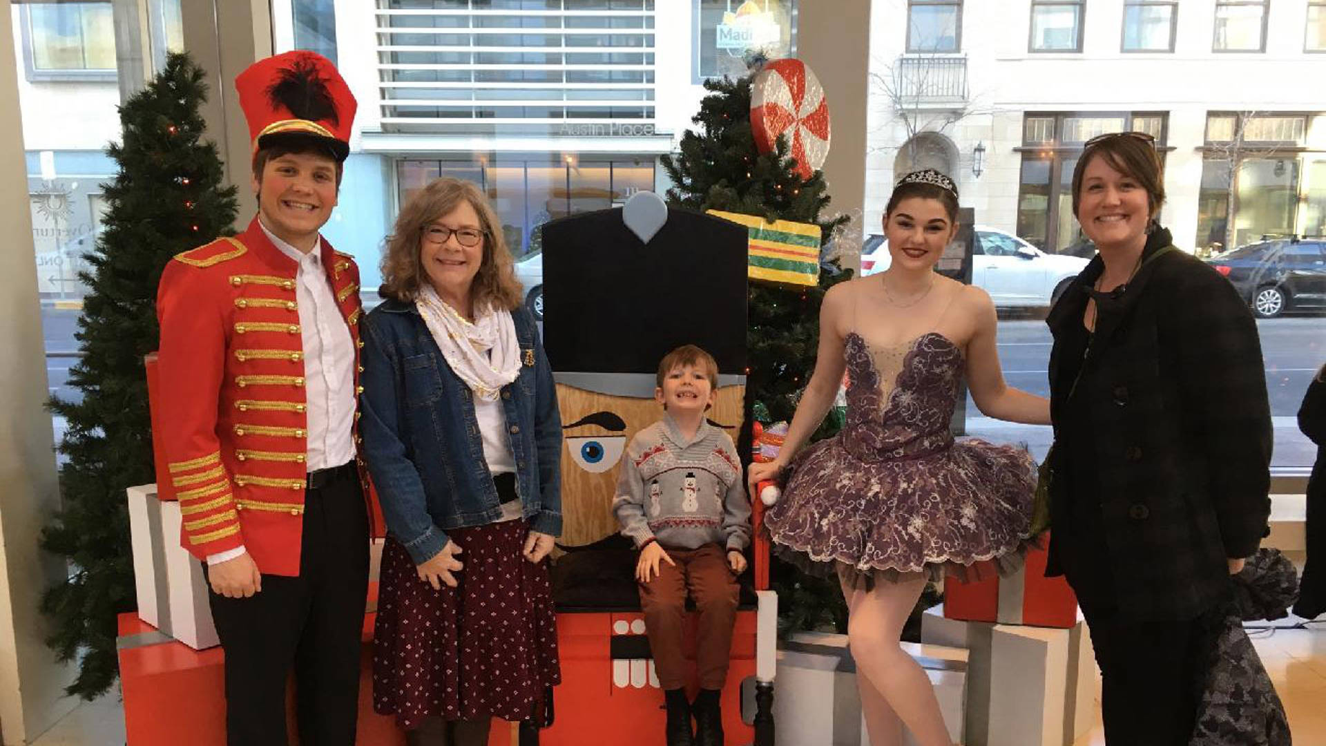 Nutcracker performers in costume pose with a family in Overture Hall Lobby in front of present boxes and trees.