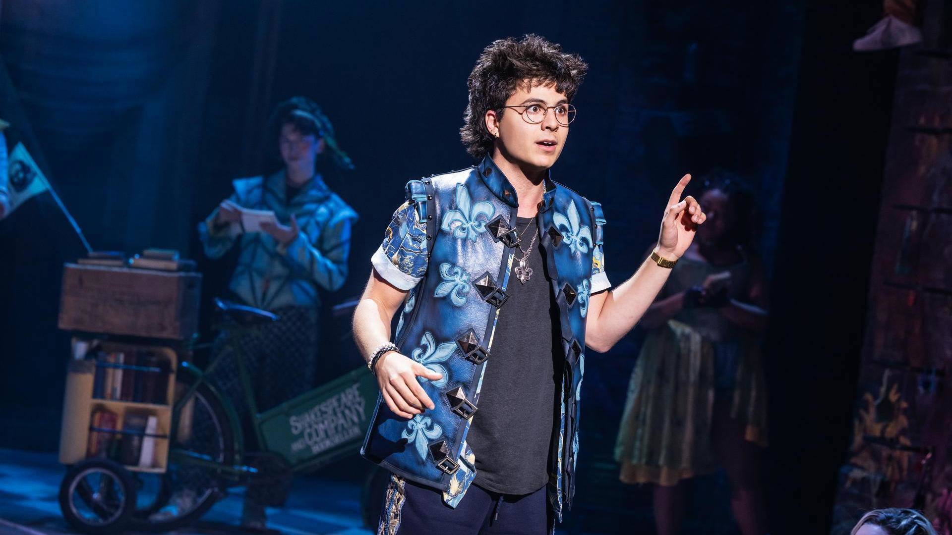 A person with light complexion, curly medium dark hair, and glasses singing with their left index finger in the air. They have a blue patterned vest on.