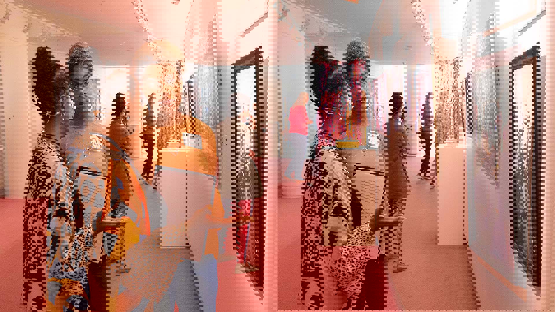 Two women with medium complections and colorful clothing stand talking to eachother in an art gallery with pink carpet.