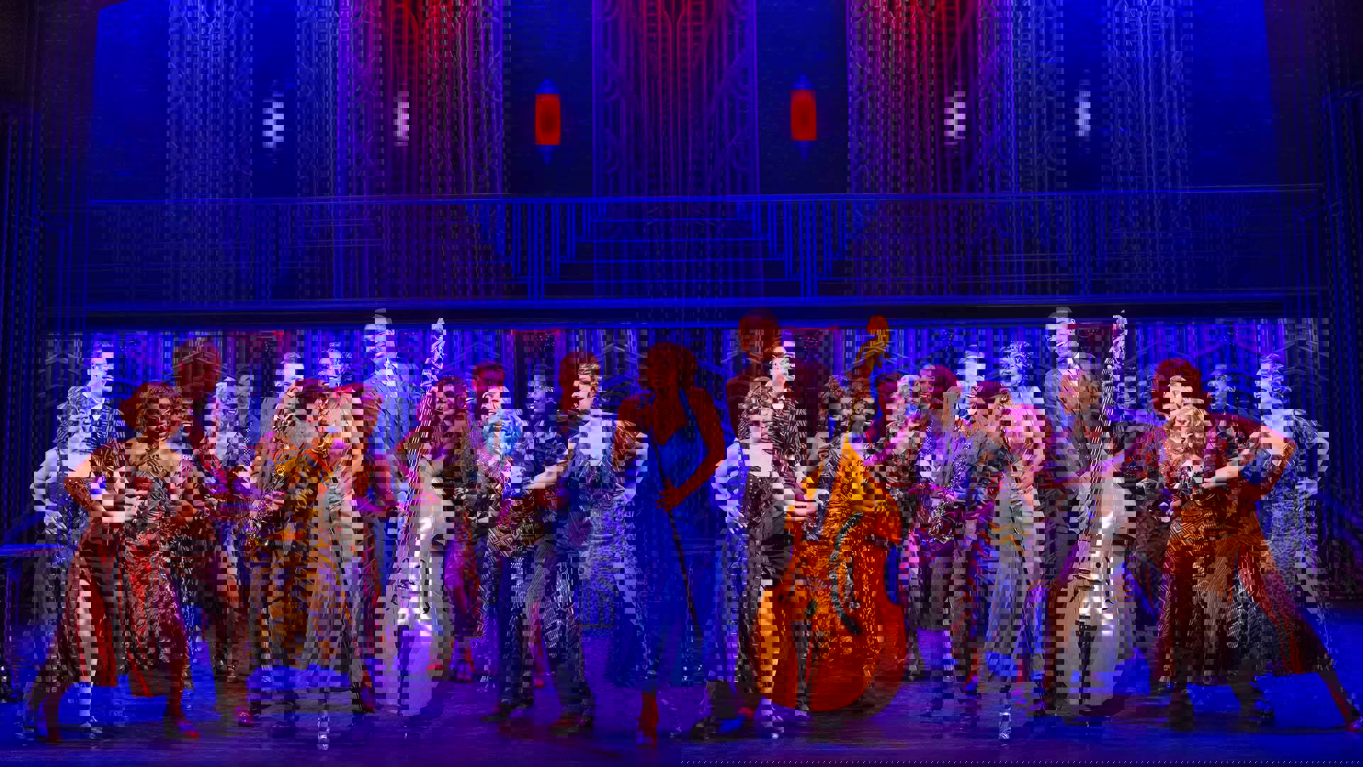 An ensemble dancing in 1930s costumes at a night club setting. Three people stand at the center performing, one holds a saxaphone wearing a blue suit, a singer with dark complexion holds a micstand wearing a blue dress and an upright bass player with dark complexion plucks while smiling.