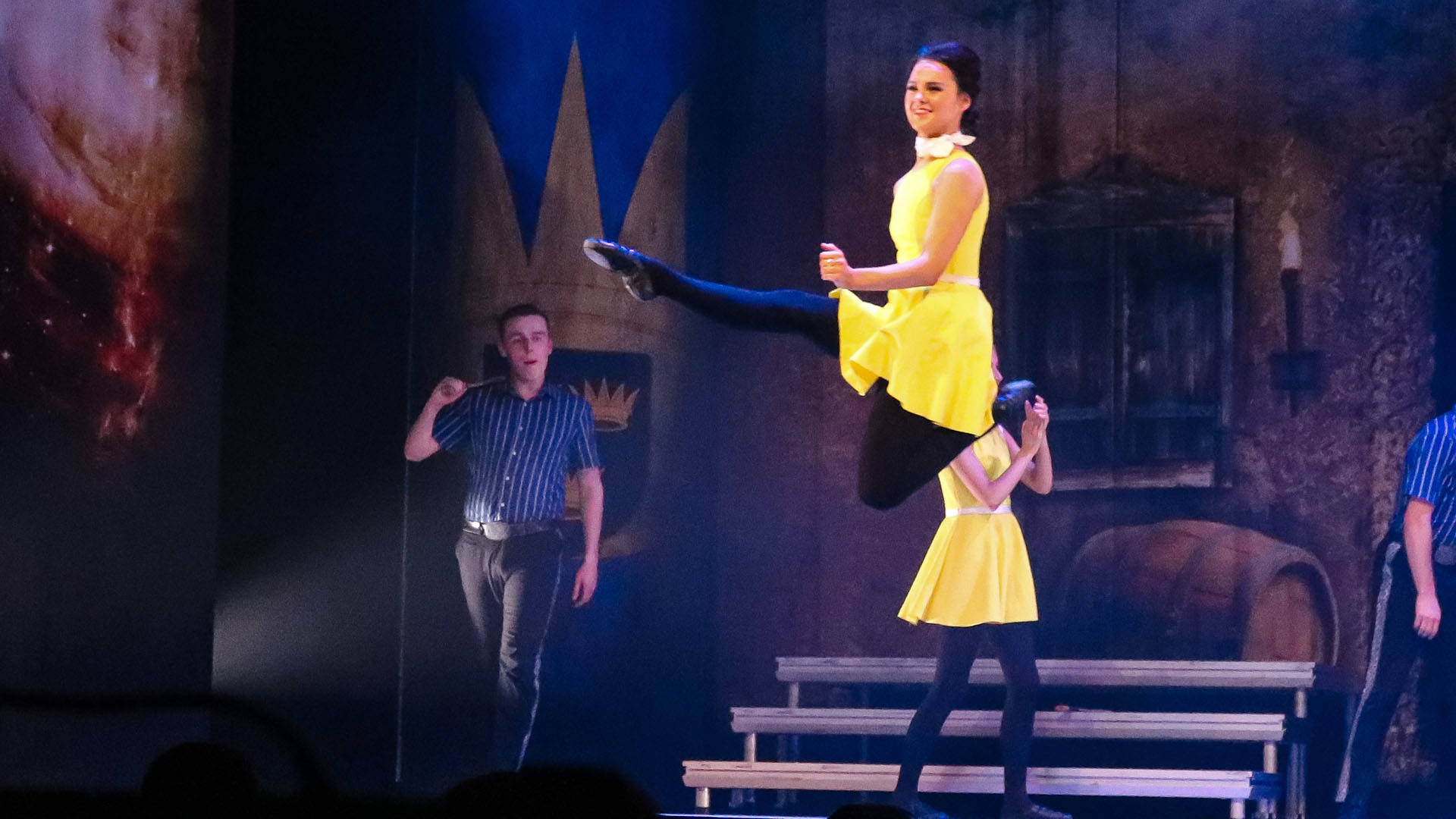 A woman in a yellow dress with dark hair  leaps into the air with her leg outstretched. Other dancers are behind her on stage.
