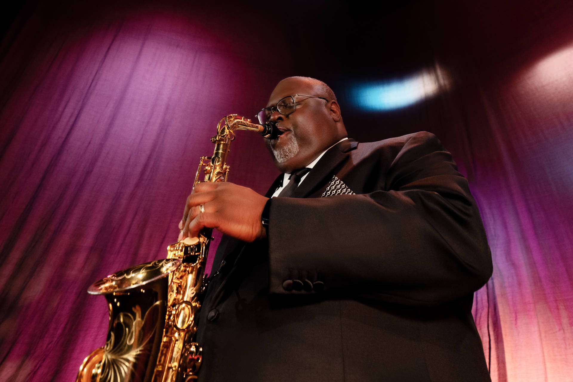 A man with dark complexion plays a saxophone in front of a pink and purple stage curtain.