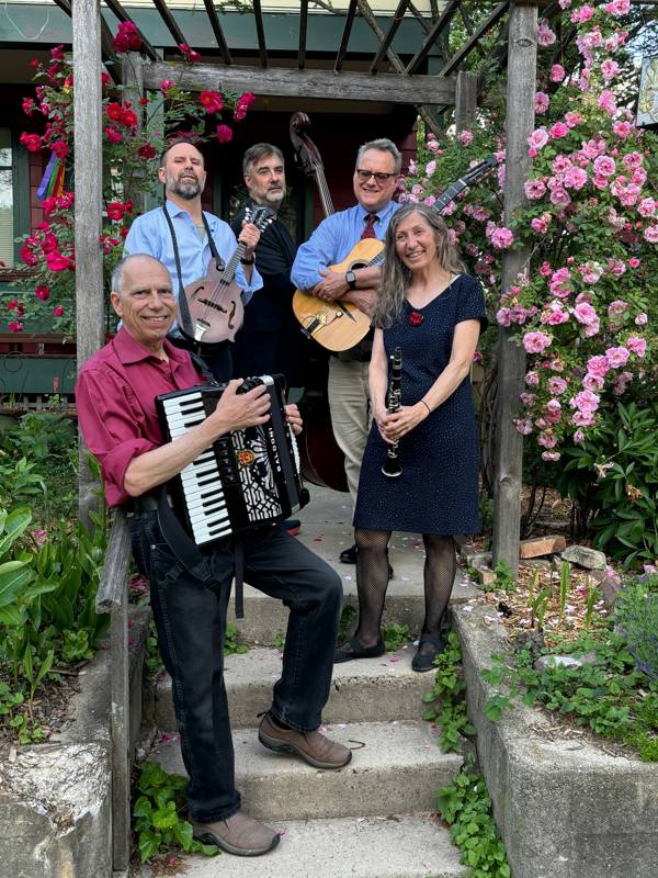 Five middle aged musicians stand outside on cement steps in front of pink flowers holding their instruments and smiling.