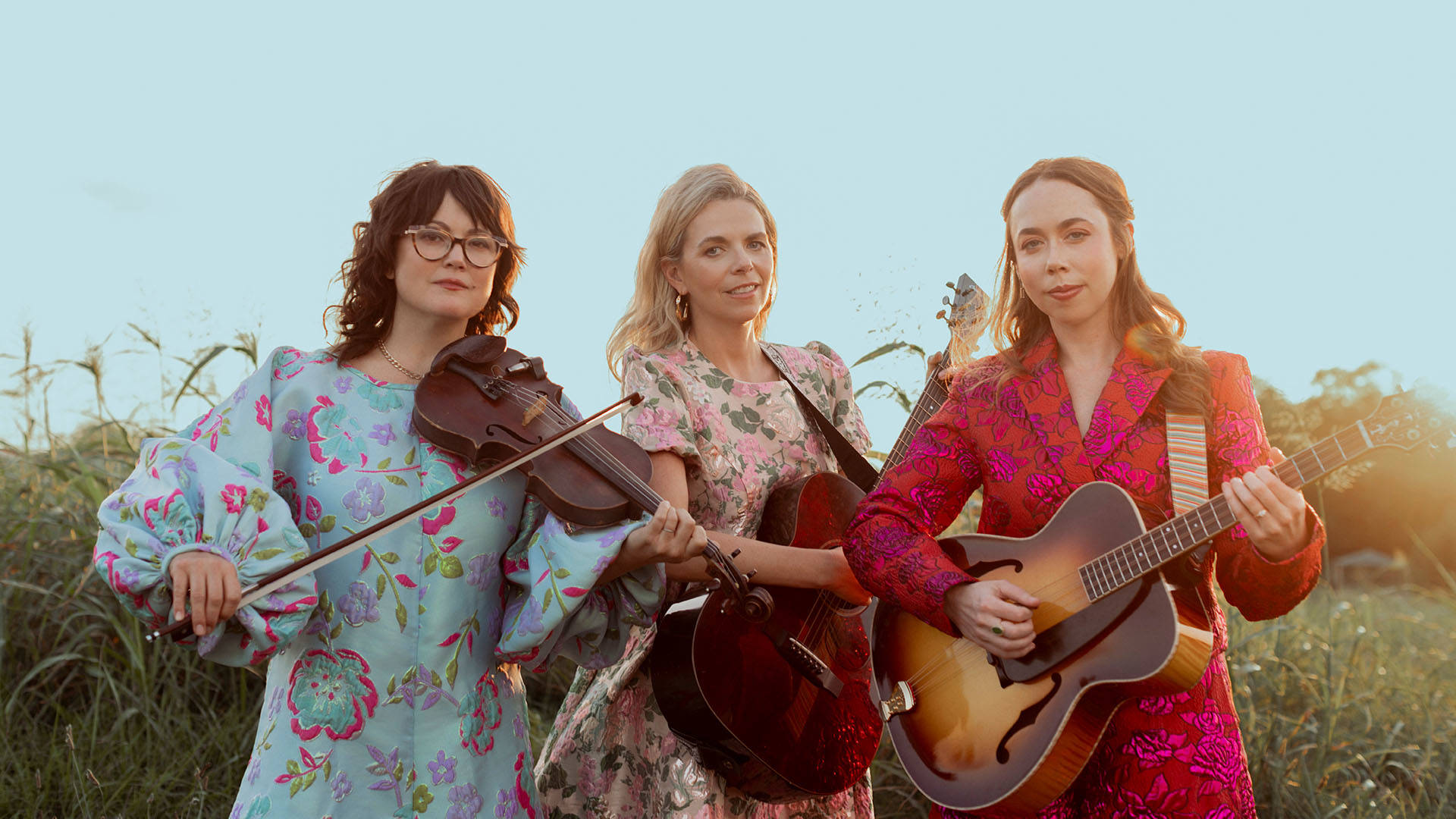 Three women with light complexions standing together in a field with wildflowers and a light blue sky. Left: A woman with shoulder-length wavy dark brown hair, dark framed cat-eye glasses, wearing a light blue and pink flowal dress with ballooned sleeves playing a violin. Center: A woman with long blonde hair in a short sleeve, pink floral dress strumming a dark wood acoustic guitar. Right: A woman with long brown hair wearing a large dark orange with pink accents blazer, holding an acoustic guitar.