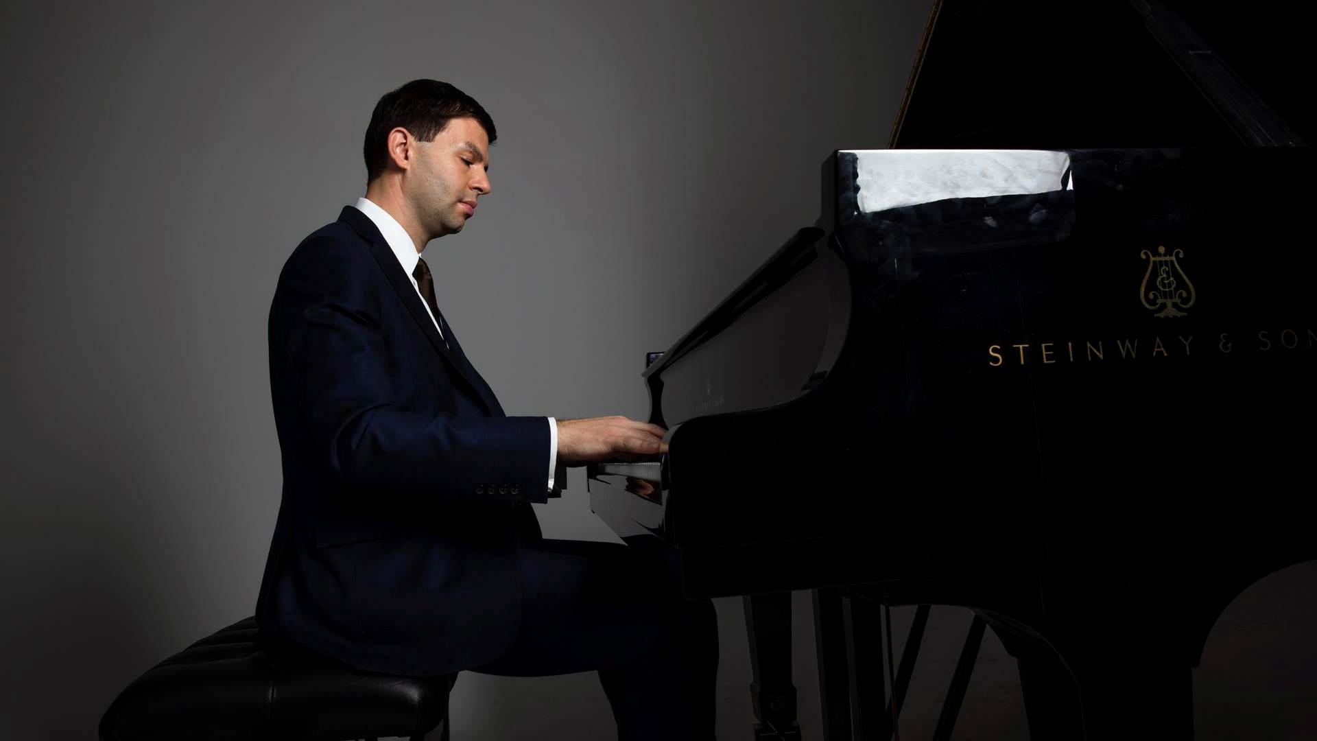 A man with medium complexion and dark hair in a blue suit playing a black grand piano against a grey background.