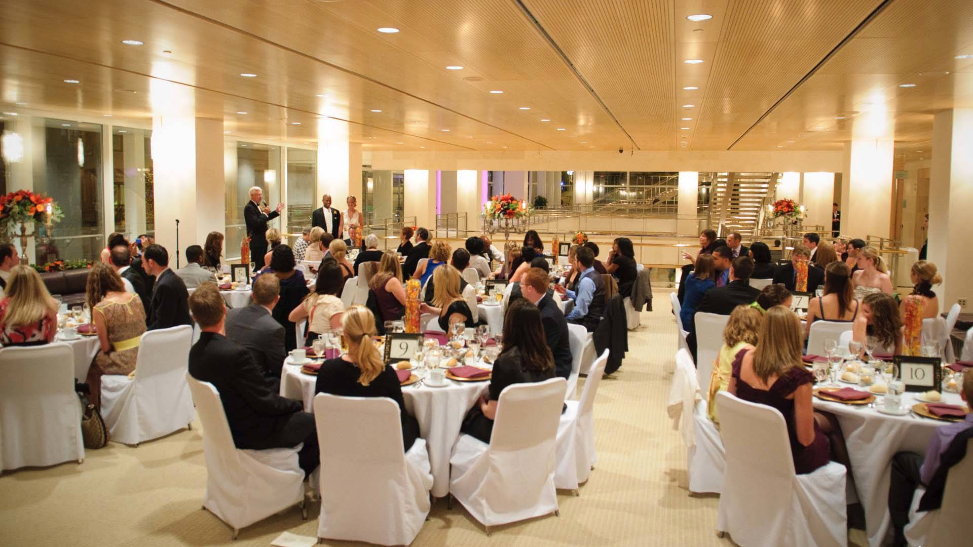 Many people sit at white tables and chairs. They are watching a person toast a bride and groom.