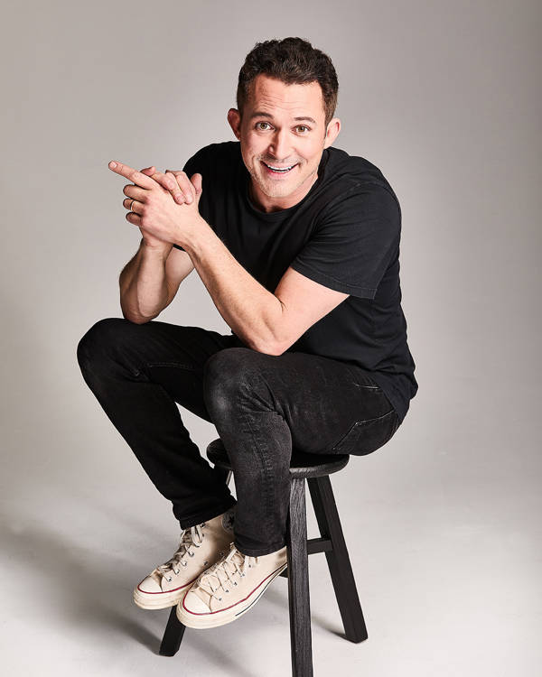 Justin Willman, a caucasian man with short brown hair and brown eyes, sitting ona black stool. He is wearing black jeans and a black tshirt. His hands are clapsed together and he is looking towards the viewer.
