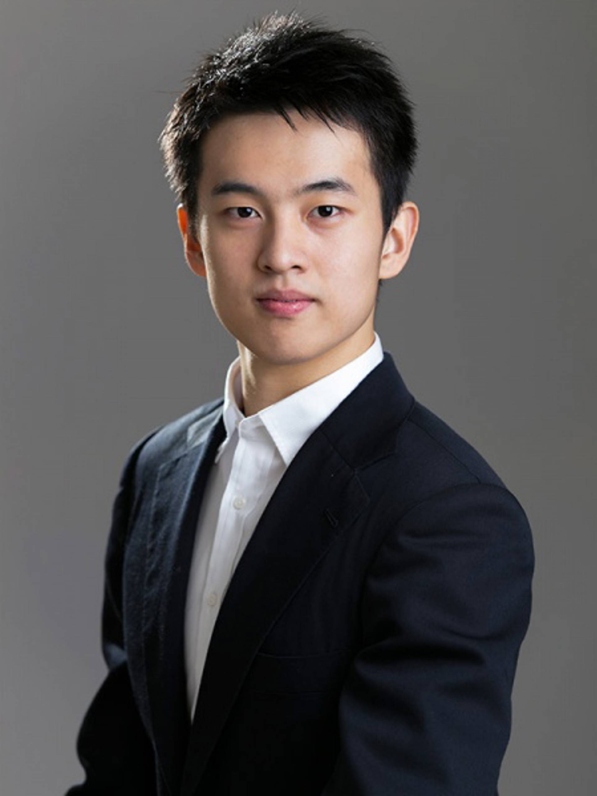 Headshot of a young man with medium complexion, short black hair, a white collared shirt and a dark jacket against a grey background