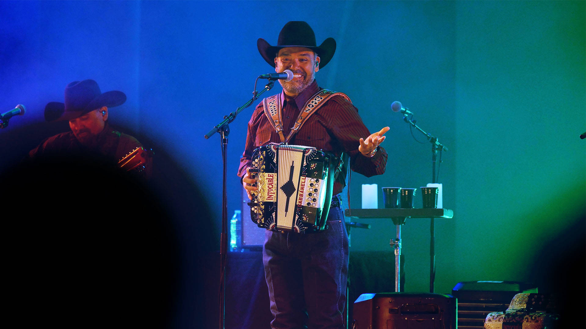 A hispanic man in a black cowboy hat, red collared shirt and jeans playing the accordion on stage.