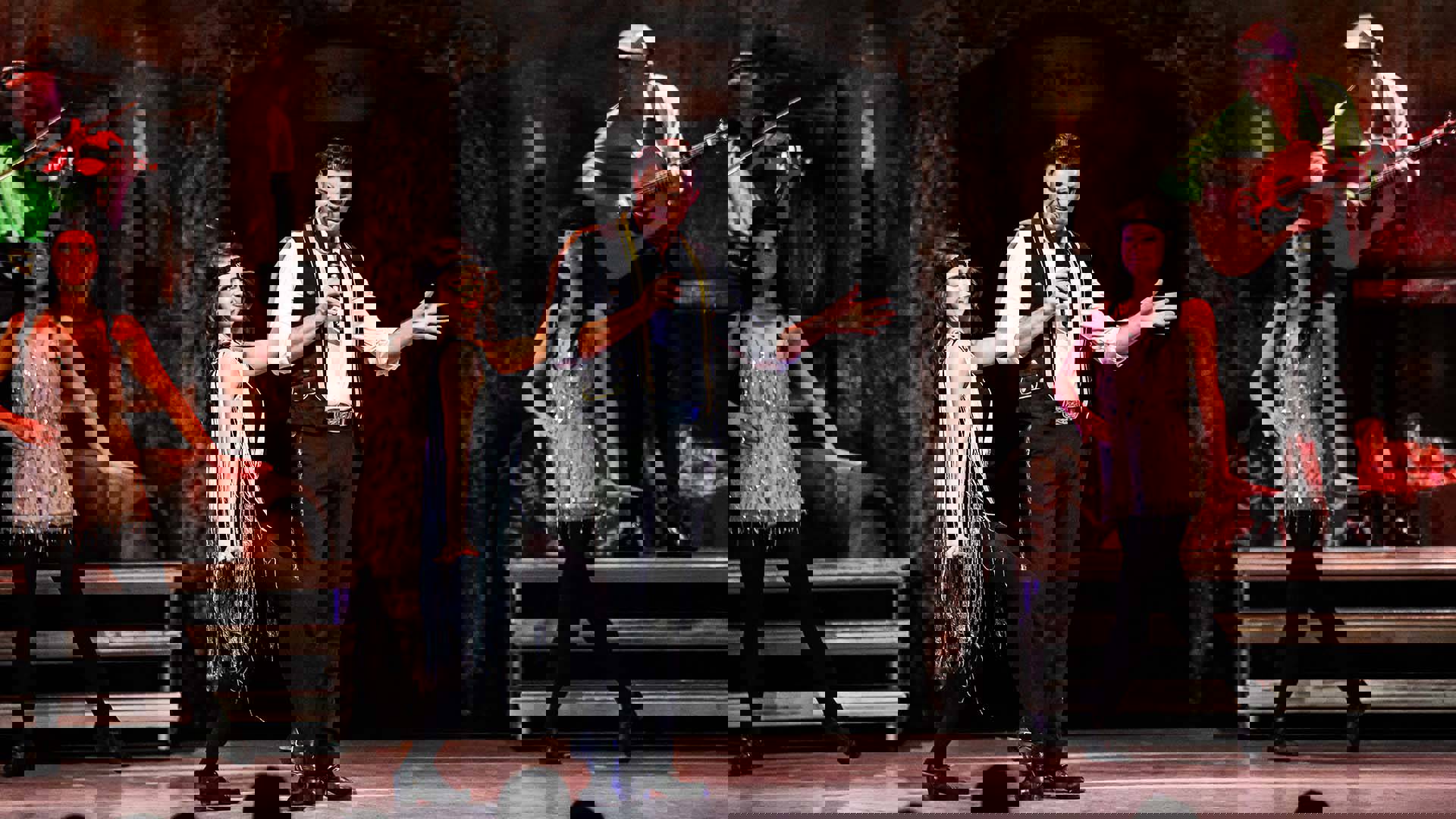 A man with brown hair, a black vest and white shirt speaks into a microphone with dancers and musicians behind him.