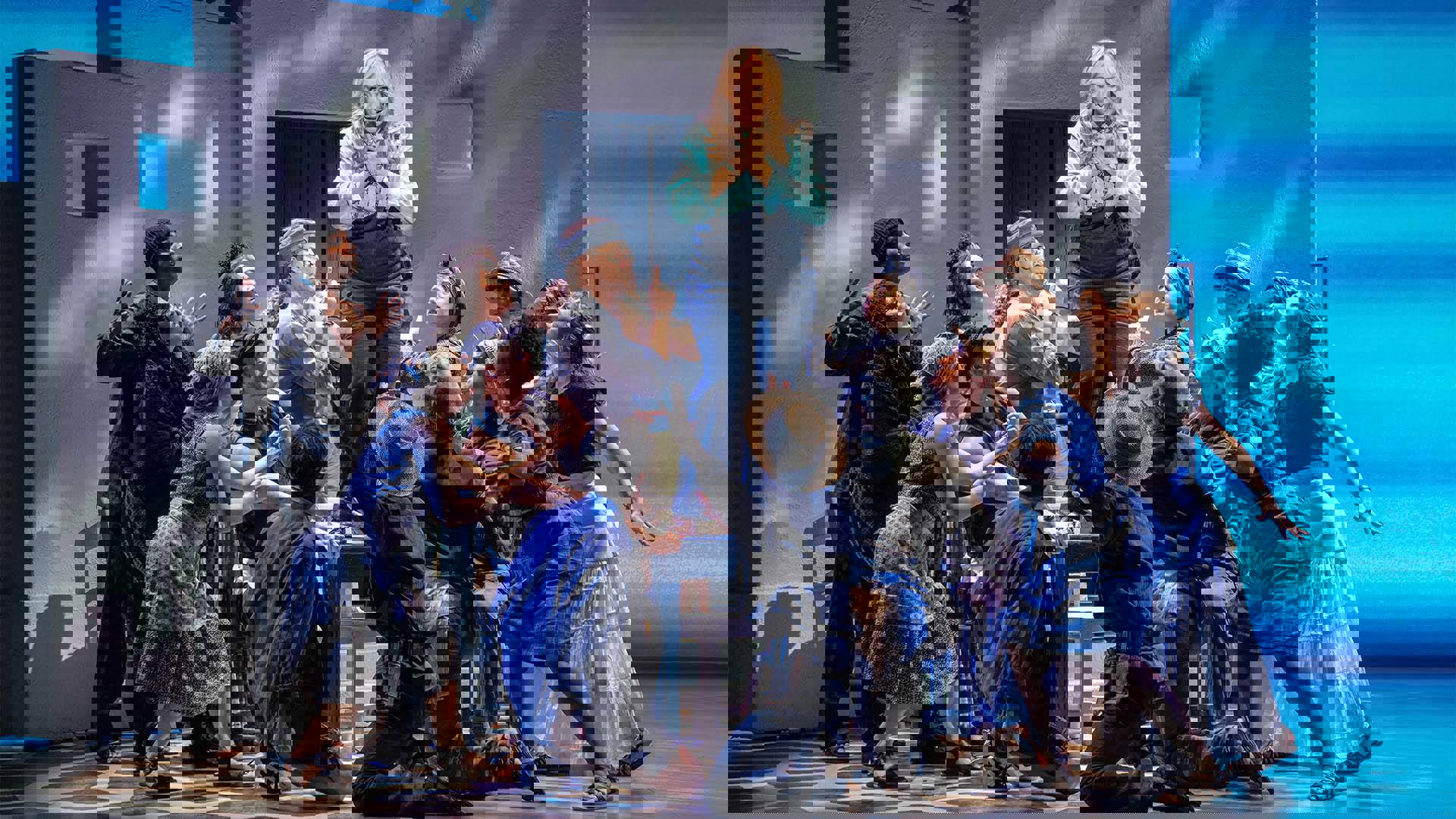 the Mamma Mia ensemble standing around an elevated woman with blond hair agains a blue and grey background.