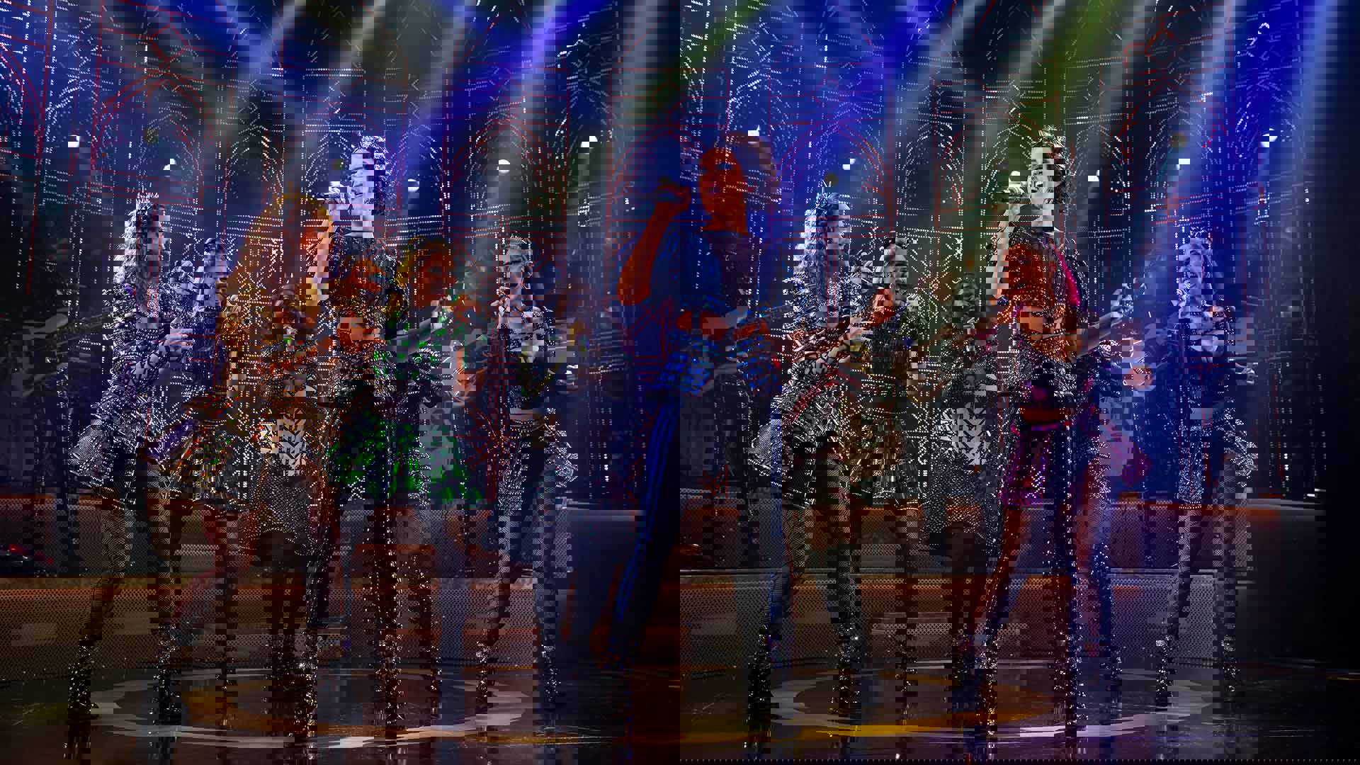 The cast of Six, in futuristic glittery outfits and holding microphones, performing on stage