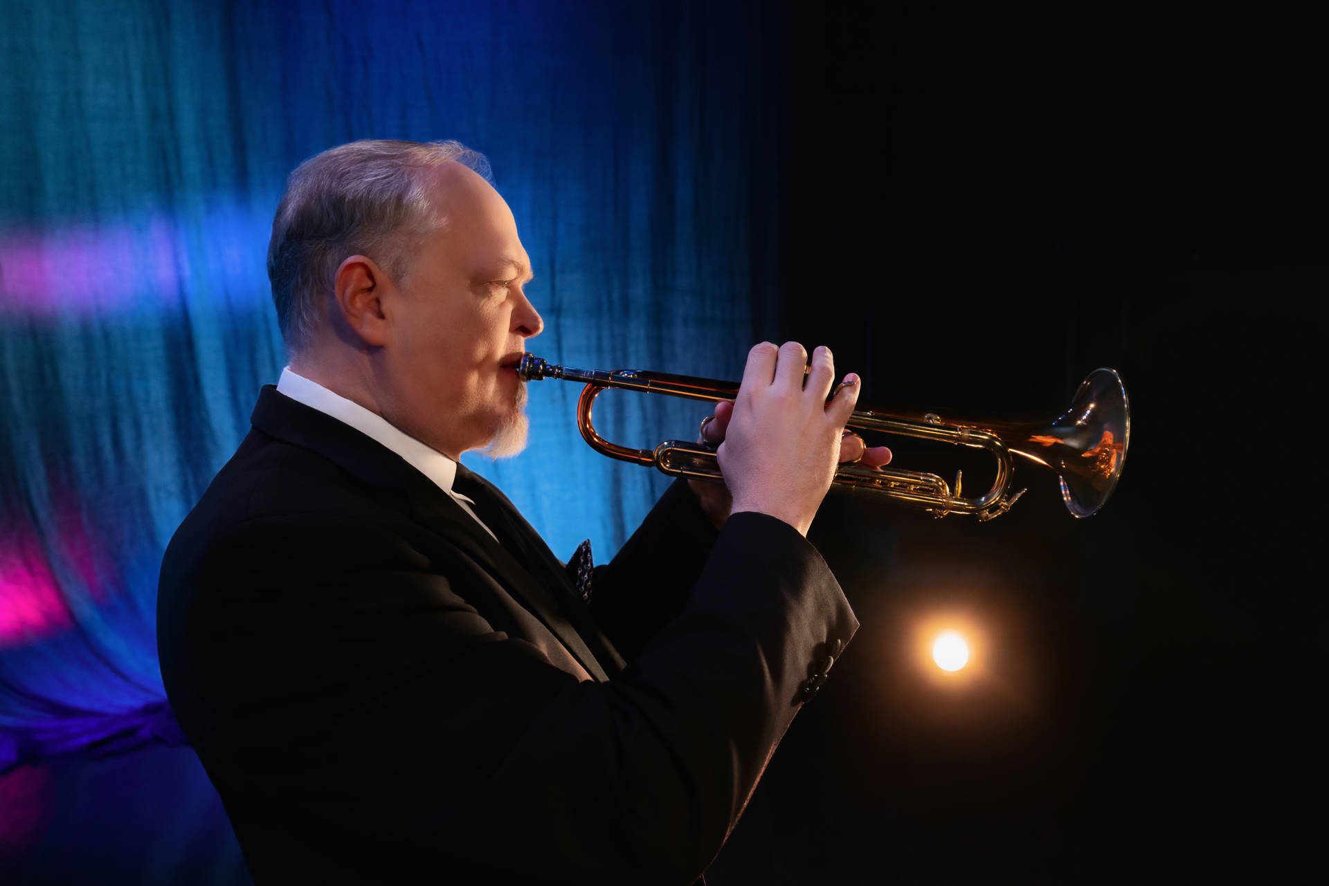 A man with light complexion and gray hair plays a trumpet in front of a blue stage curtain.