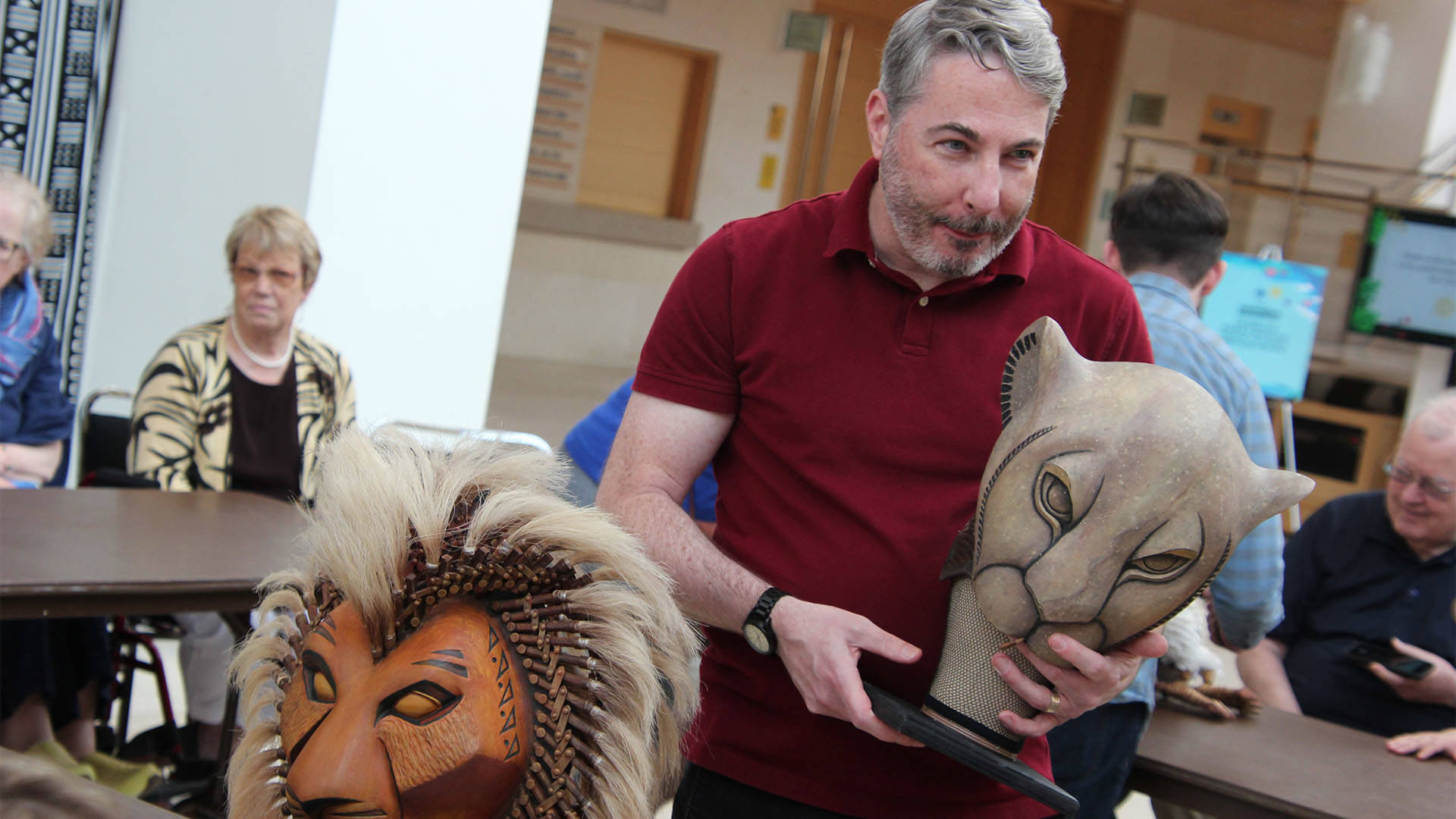 A man in a red shirt and grey hair holds a beige lioness mask.