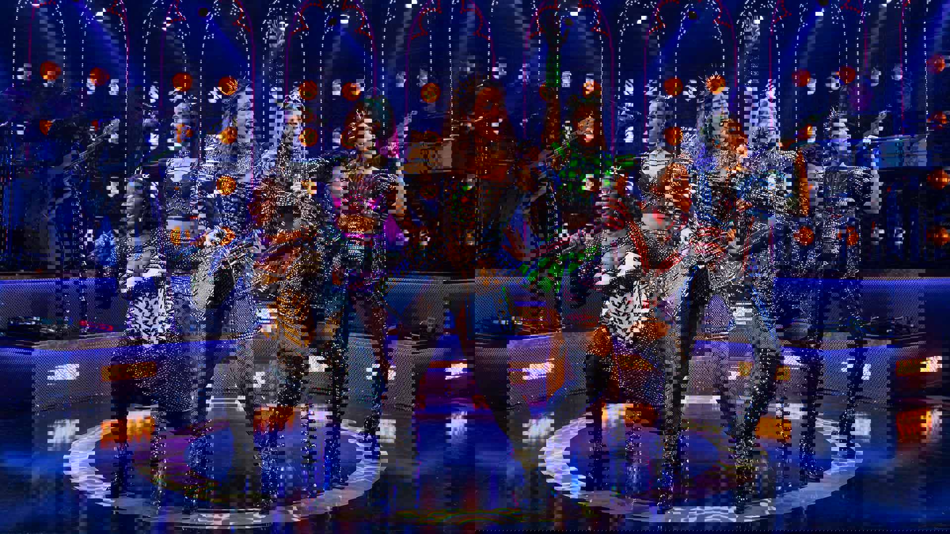 The cast of Six, in futuristic glittery outfits and holding microphones, performing on stage