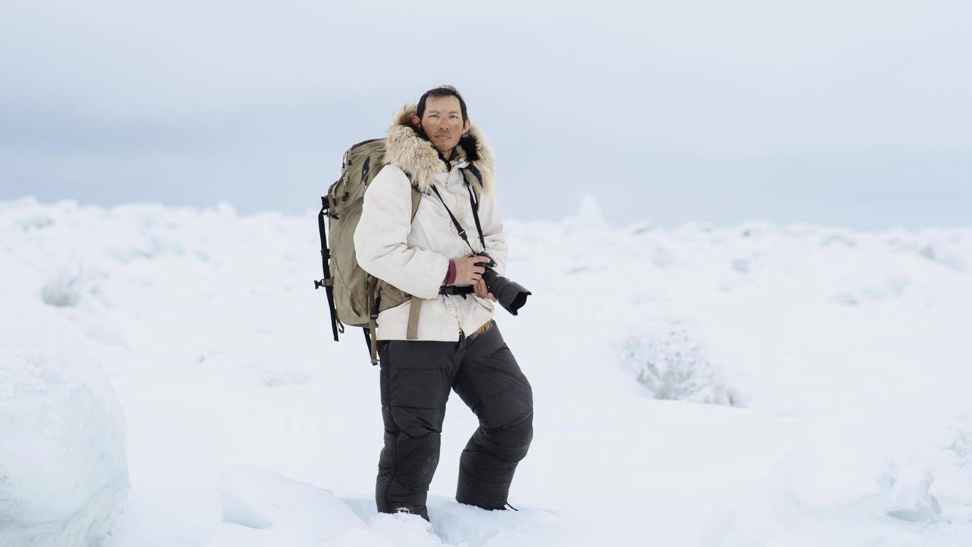 Photographer Kiliii Yuyan stands in snow holding a black camera. He is wearing black snow pants and a white coat with a tan backpack on his back.