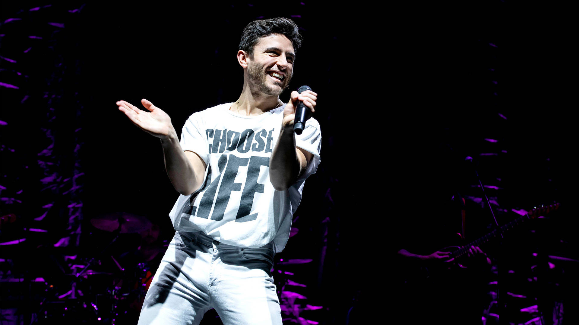 A white man with short brown hair smiles and is clapping his hands as he performs. He is wearing all white and his shirt has the text "Choose Life"