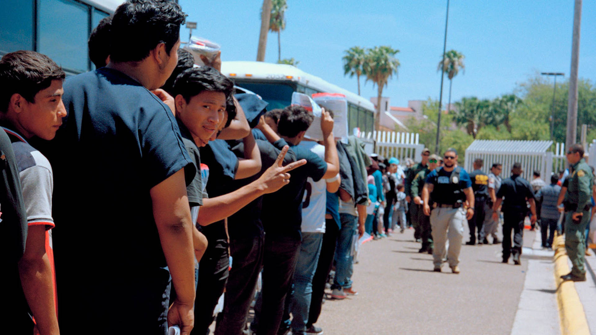 A line of young men mostly facing away, while an officer walks along looking towards them. One man in line has his head turned back towards the camera, and is holding up a peace sign with his fingers.