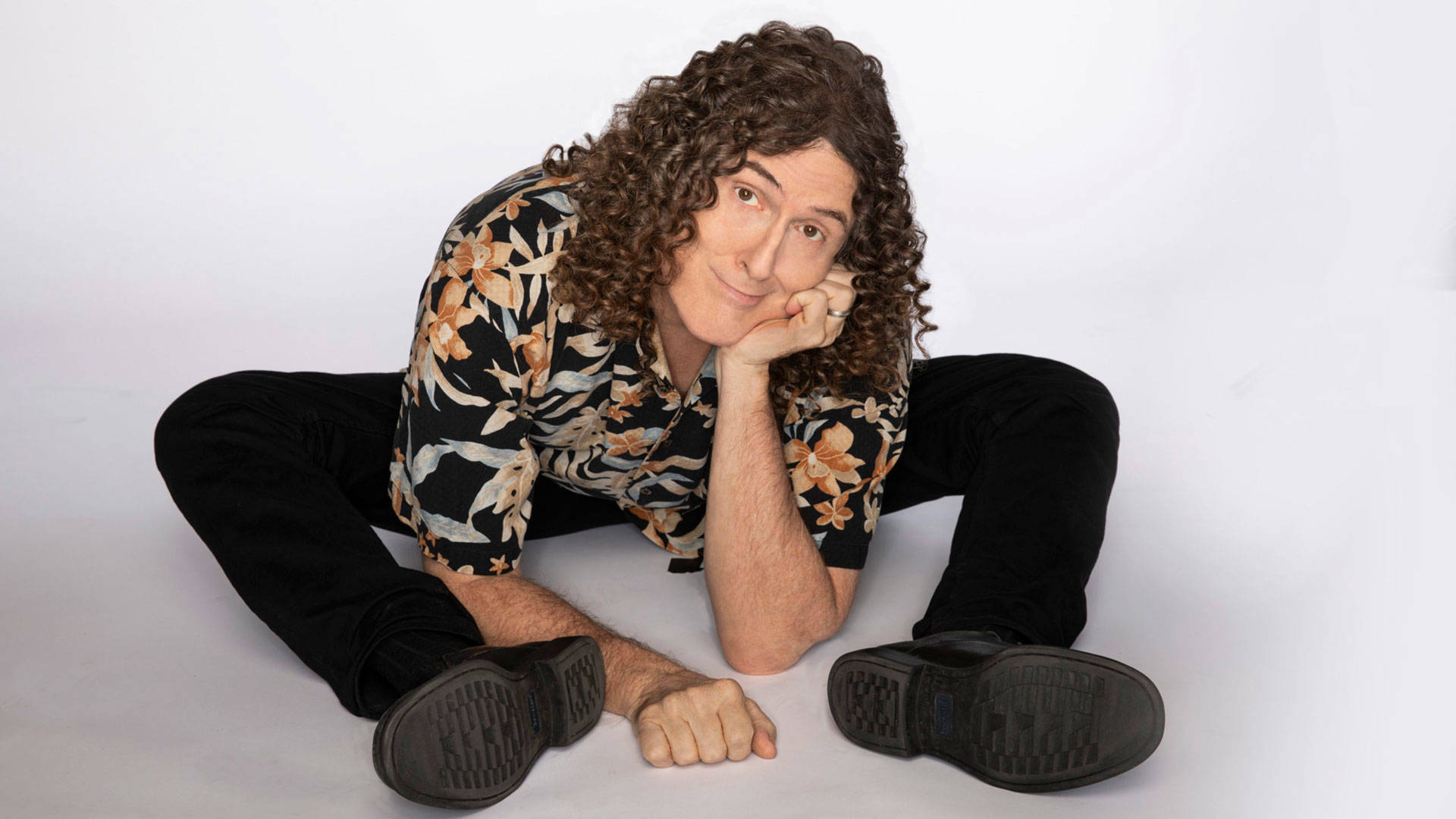 "Weird Al" Yankovic sits on the the ground on the ground and smiles towards the viewer. He has iconic long curly hair and is wearing a floral shirt. He is resting his head in his hand