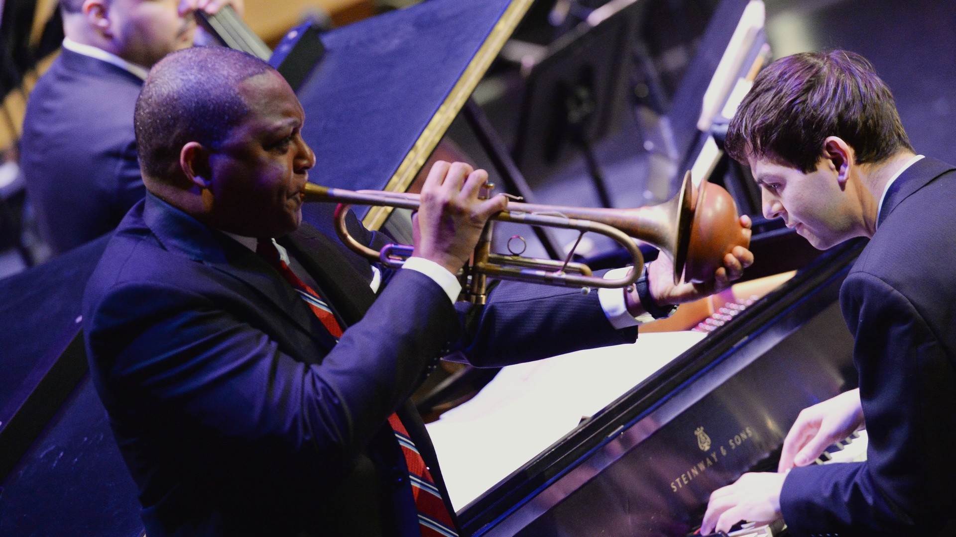 A man with dark complexion playing trumpet and using a mute. A man with light complexion and brown hair is playing piano behind him.