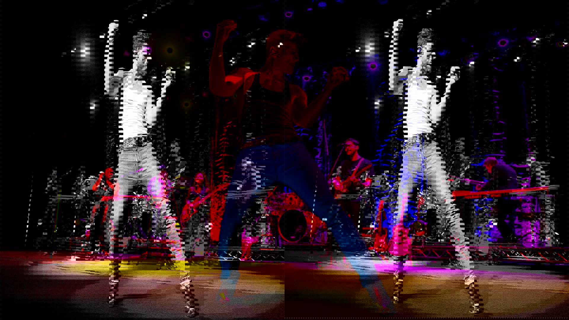 A white man in jeans and a white tank top dancing on stage holding a microphone.