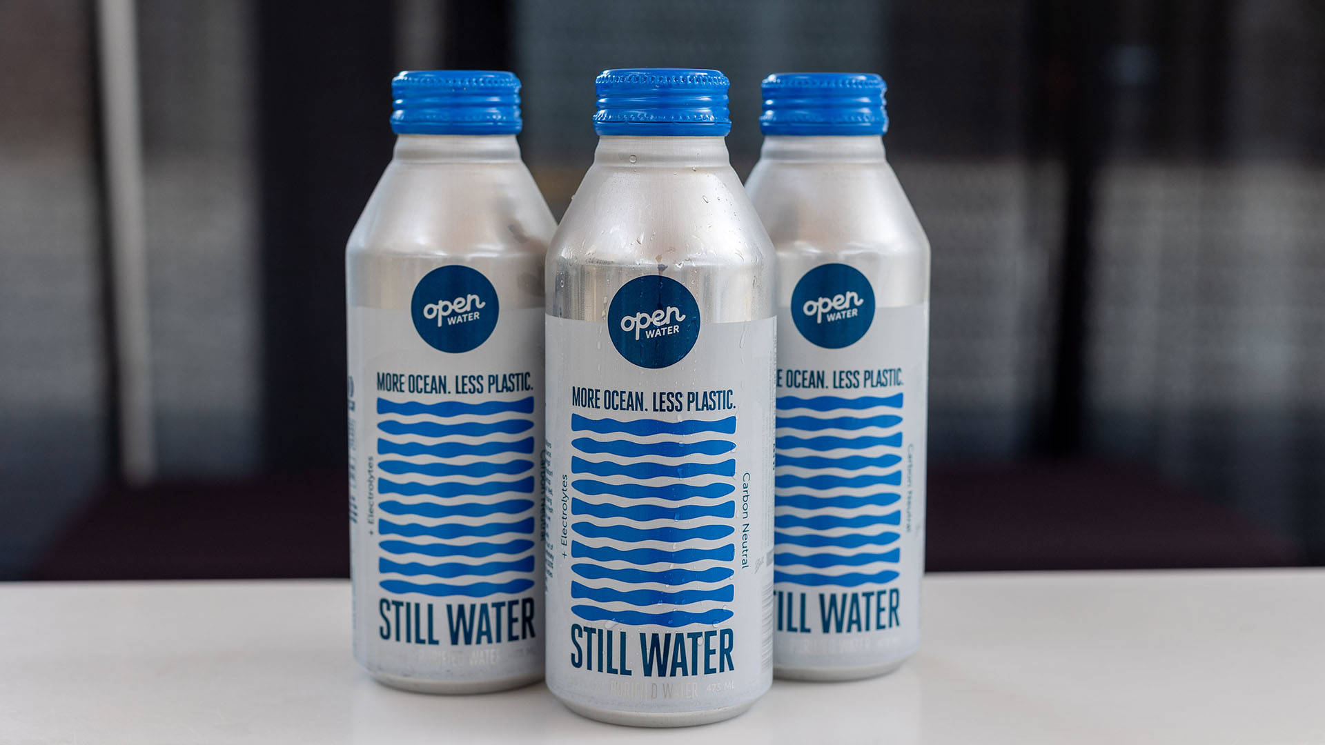 Three aluminum water bottles with blue caps that say "Still Water. More Ocean, Less Plastic."