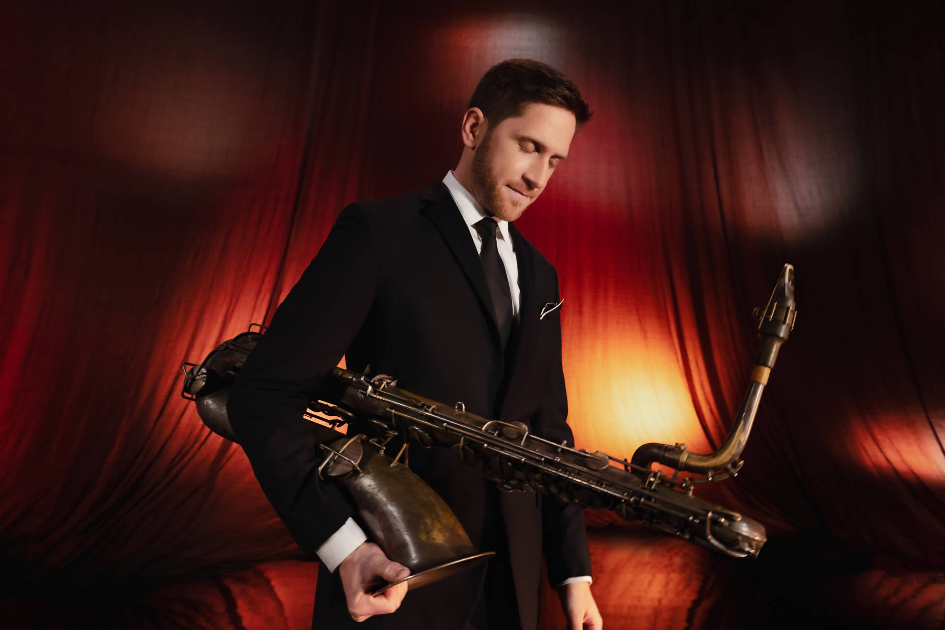 A man with light complexion and brown hair holds a saxophone and looks downward in front of an orange stage curtain.