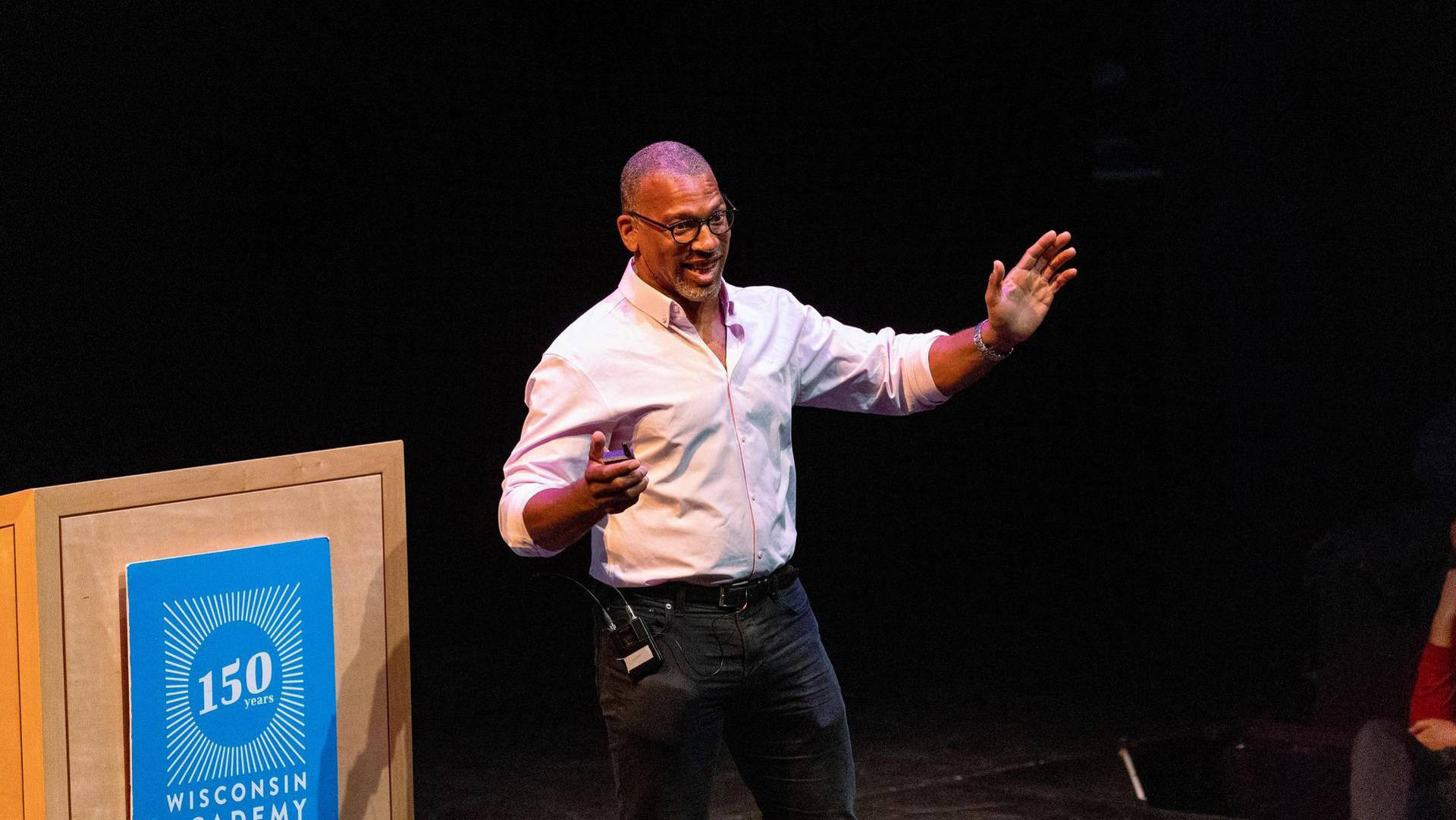 An african american man with buzzed greyish hair, glasses, and a white button-up shirt speaking next to a lecturn
