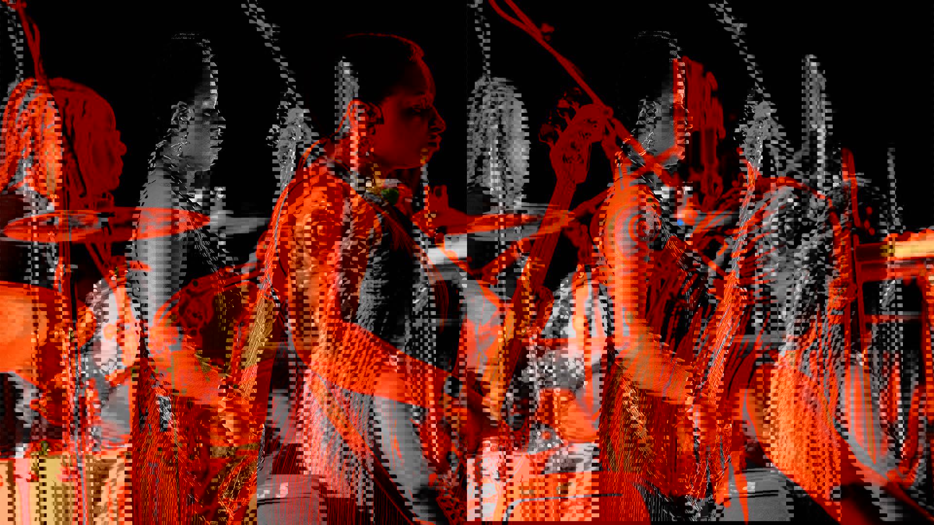 A woman with dark complexion and short dark hair in a dark top playing an electric bass. She is cast in orange light