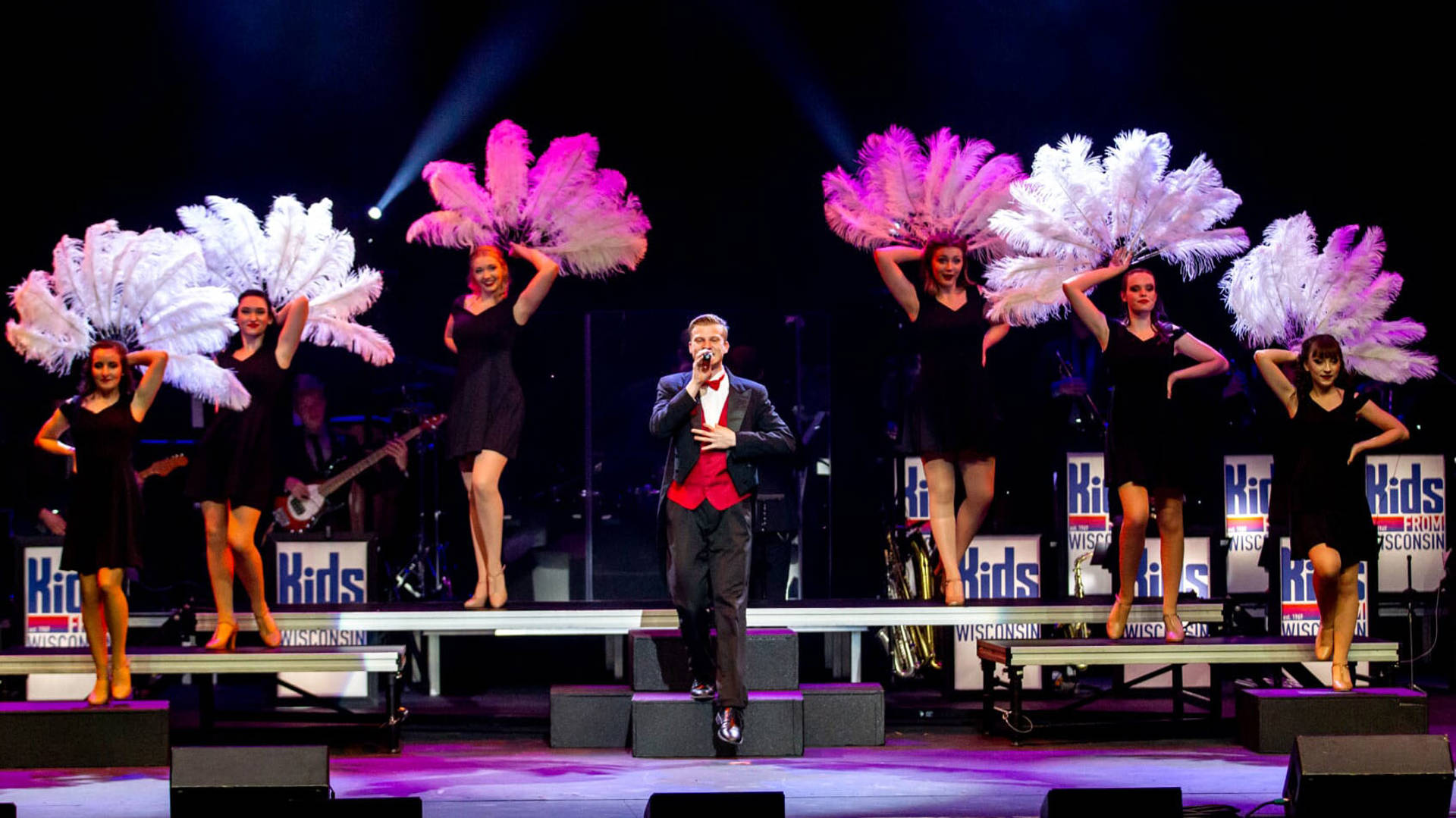 Kids from Wisconsin performing on stage. One performer is at the center in a tuxedo with red vest and six behind in black dresses holding up larger feathery fans.