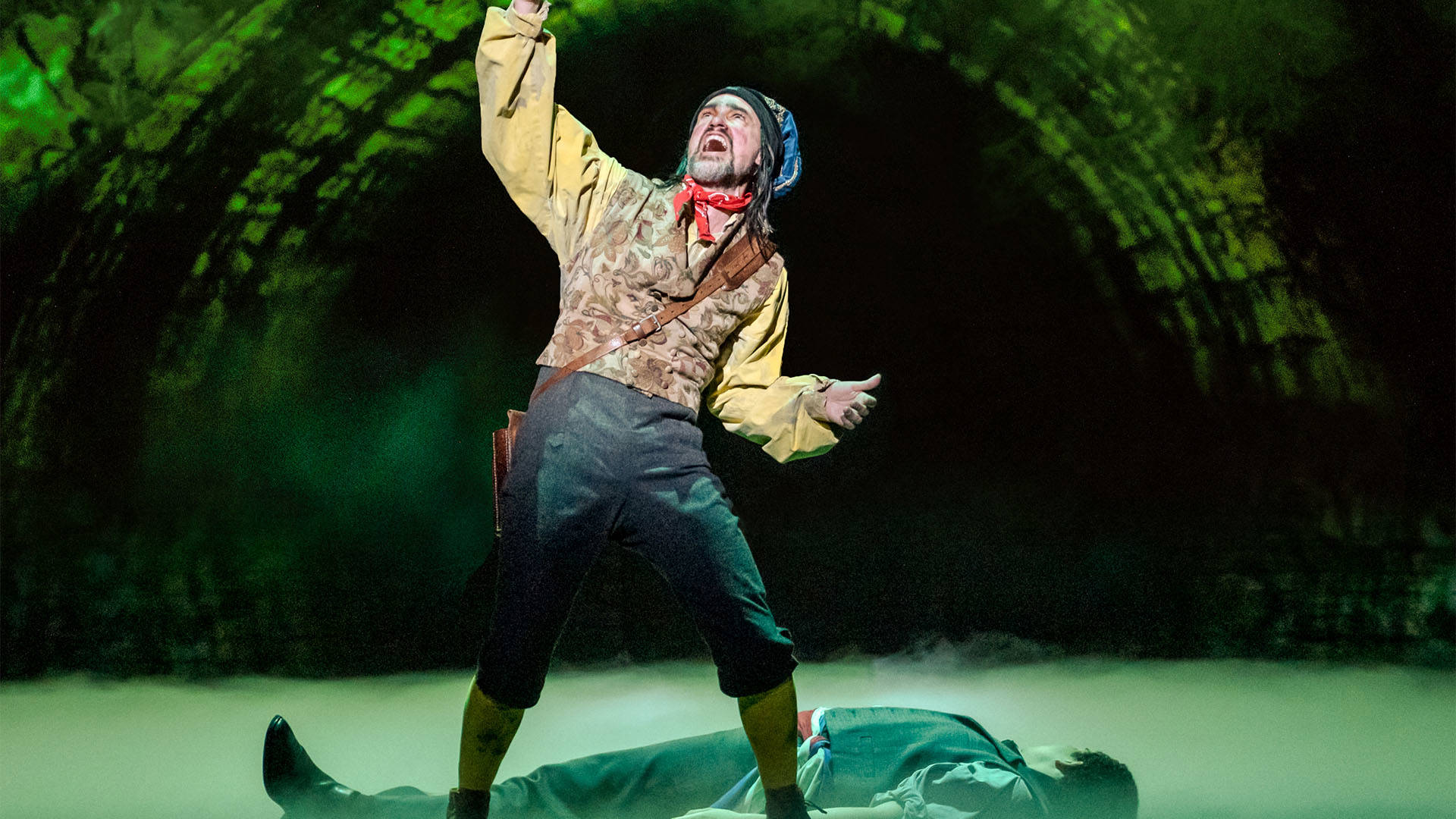 A man in a yellow top and red hankerchief sings over another man lying motionless on the ground.